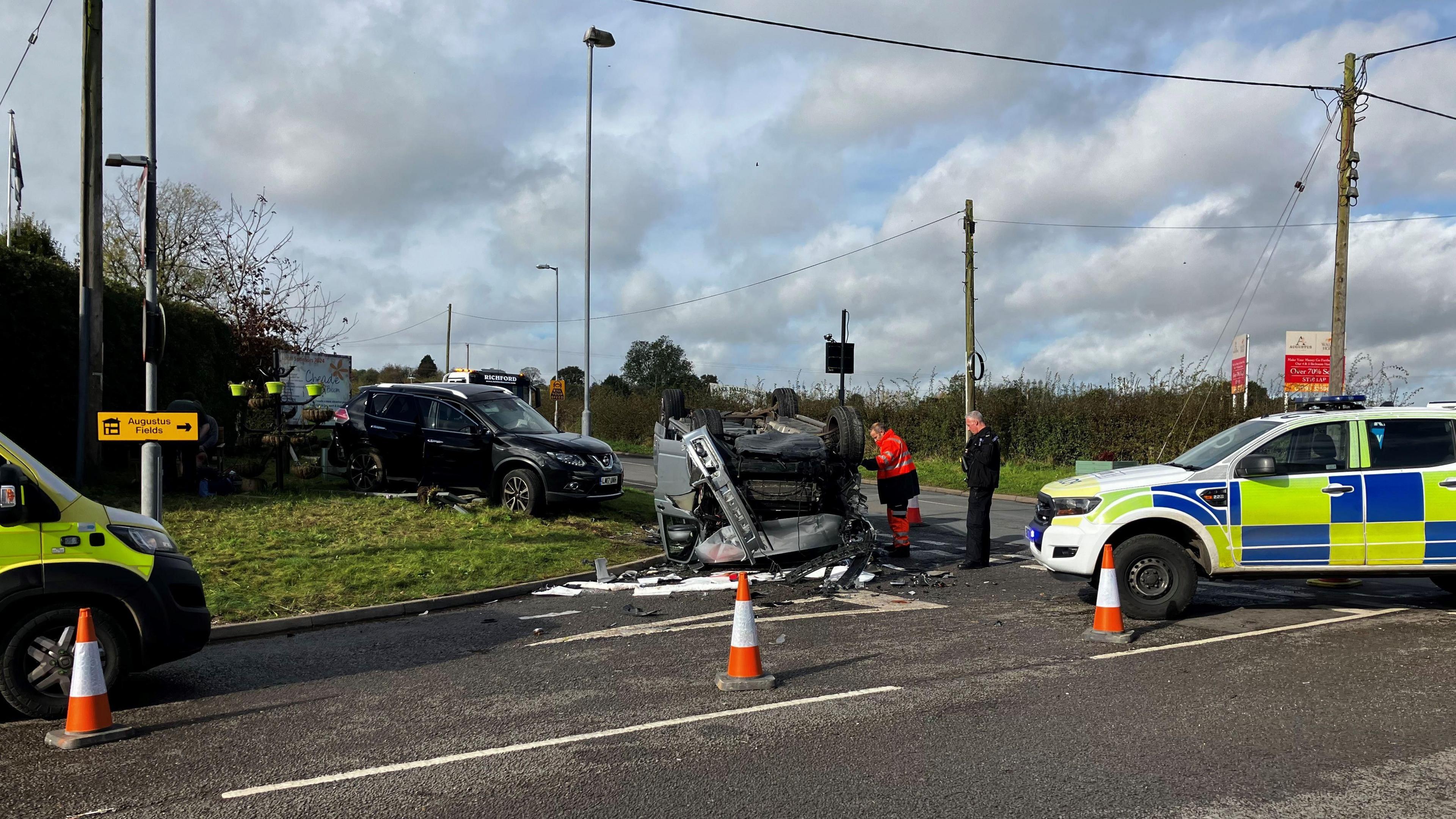 A police car and ambulance are parked nearby an upturned vehicle which is being attended by emergency service personnel.