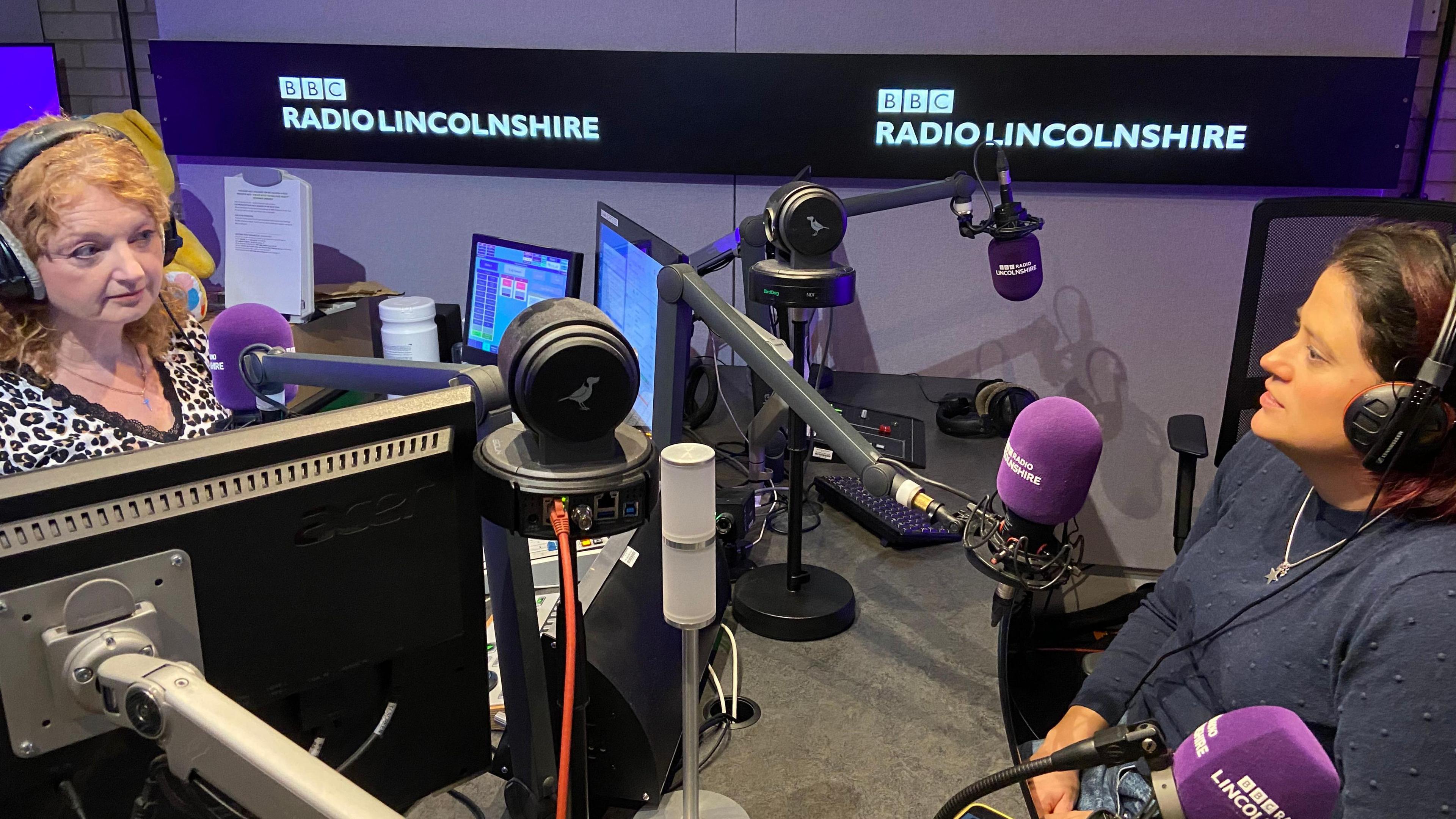 Interior of radio studio with Frances Finn presenting surrounded by monitors and technical equipment. Regan Harris is looking at Fran from the left. 