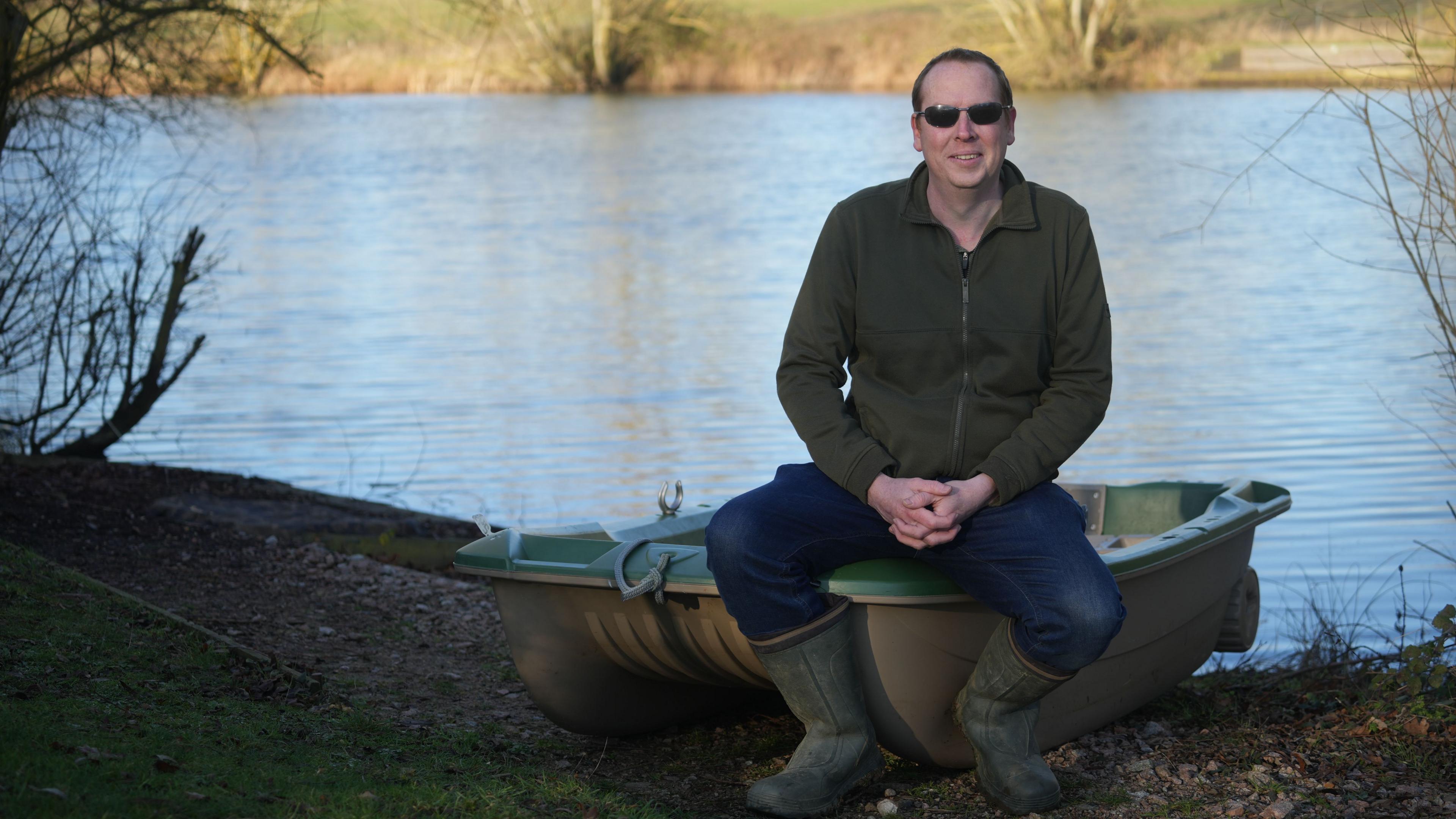 Robert Chandler sitting on the edge of a fishing boat which is on dry land, close to a body of water. Mr Chandler is wearing dark sunglasses, a green jacket, blue jeans and green wellington boots.