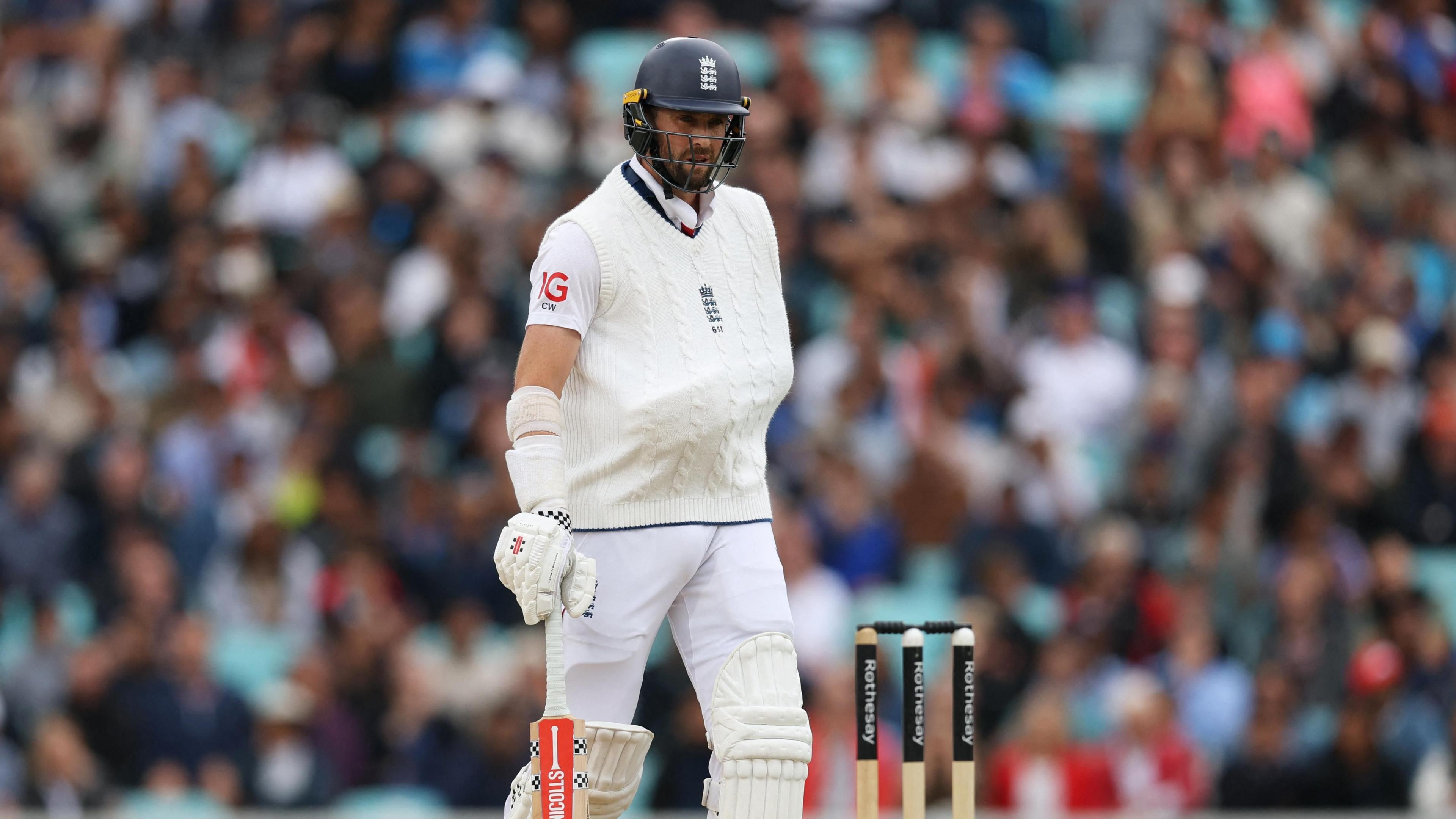 A man wearing a black helmet and cricket whites is standing next to wickets, with a bat in his hand. He has one arm strapped to his body and tucked under a cream knitted sweater vest.