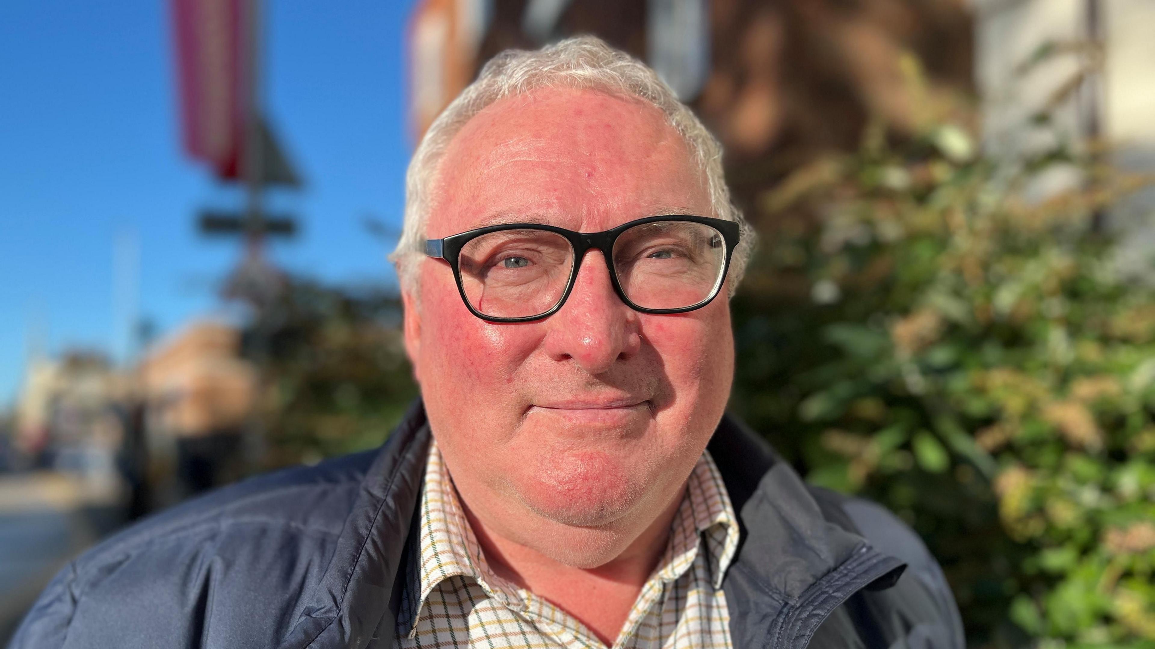 A man around the age of sixty stands close to the camera in front of a hedge and blue sky, slightly smiling with his mouth closed. He has grey, short hair and wears thick rimmed black glasses. He wears a checked white shirt and a navy jacket.