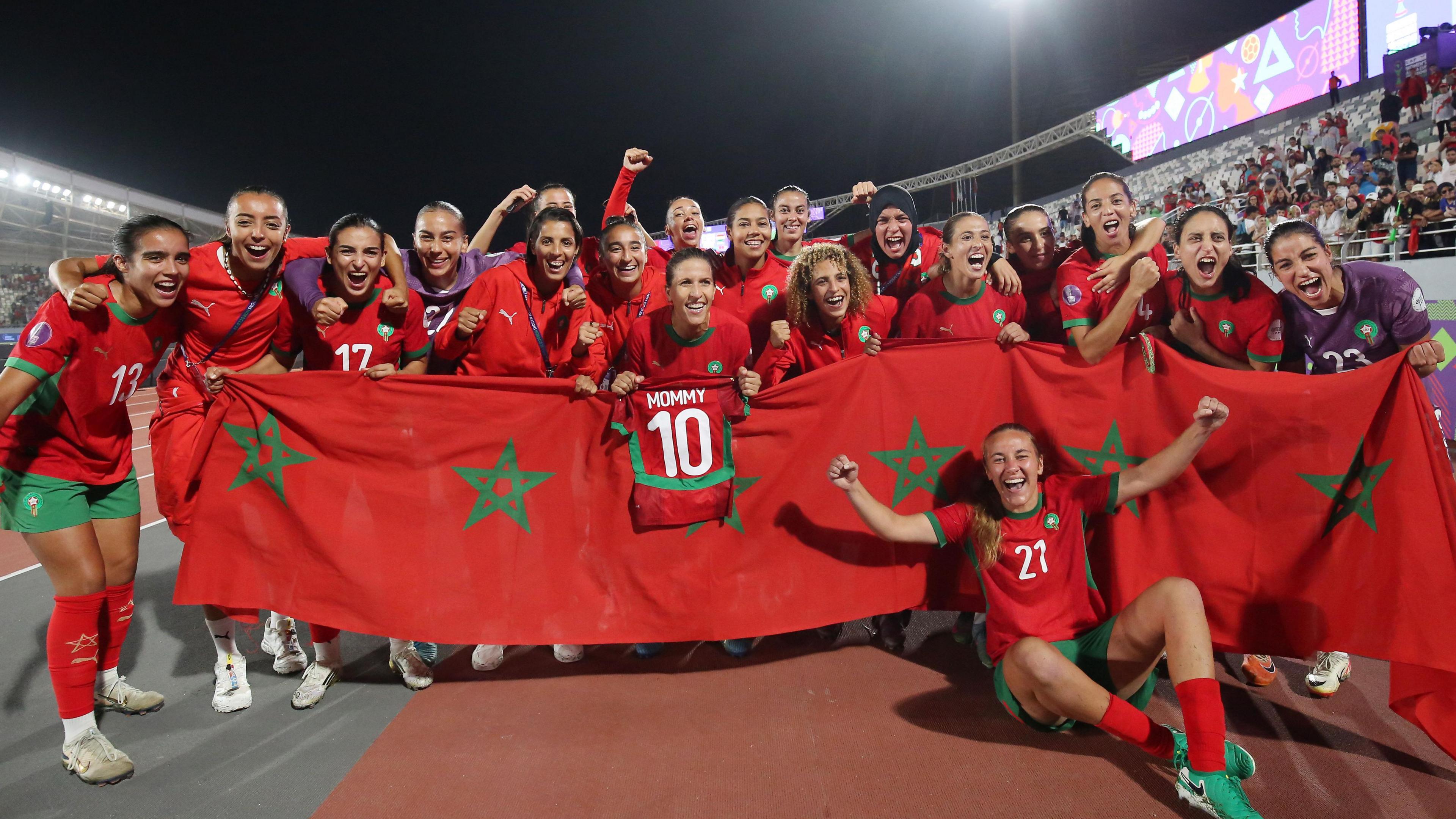 Morocco players celebrate inside a stadium after winning their Women's Africa Cup of Nations semi-final by gathering around a large red banner made up of several Moroccan flags joined together, with most players standing behind it, while one of them hangs a mini kit over the top that shows the number 10 and the word mommy on its back