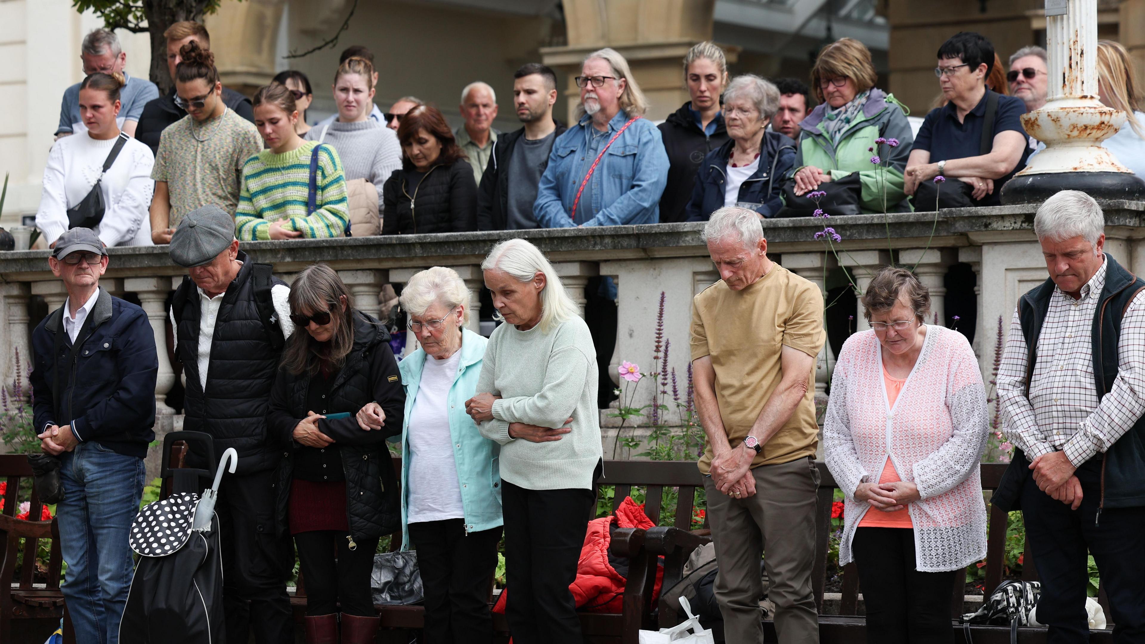 People of all ages in various kinds of casual attires stand in silence some with their heads bowed and arms folded