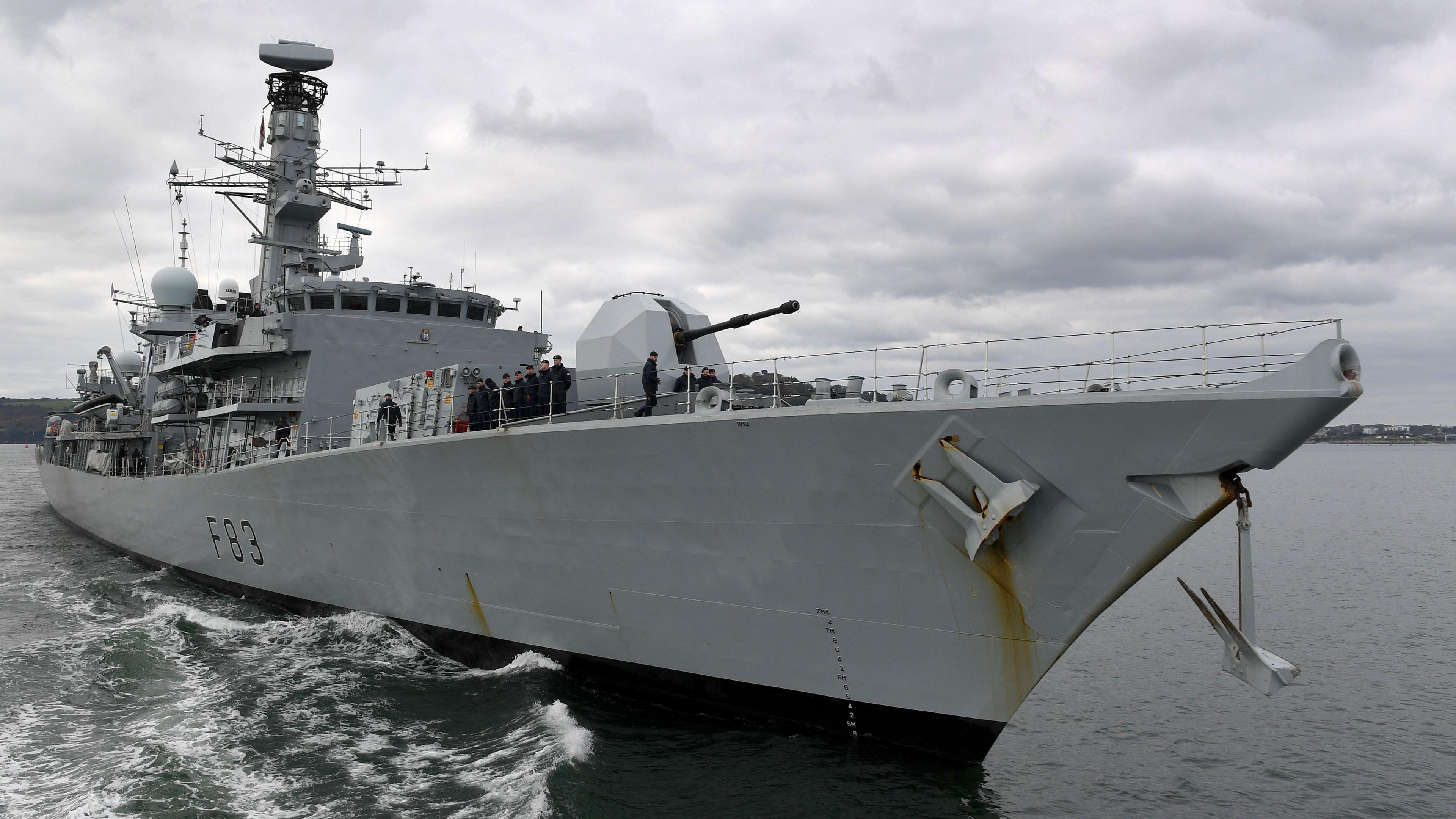 Members of the crew of HMS St Albans standing on its deck. The warship, seen here sailing, is painted light grey and has the hull number F83. It is equipped with various antennae, radar systems, and a large gun turret on the deck. The background shows a cloudy sky and a coastline in the distance. 