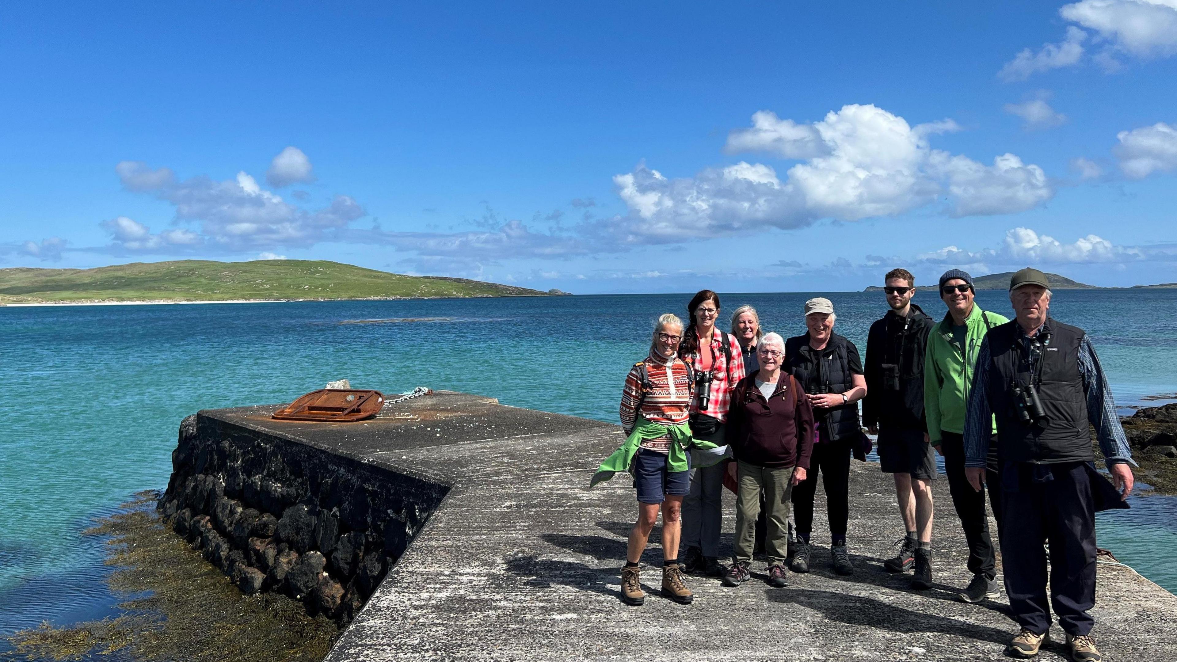 Ochdnar nan seasamh air cidhe ann am Barraigh air là brèagha samhraidh. Tha a' mhuir gorm agus ciùin, cnoc beag gorm air an cùl agus an speur gorm 's chan eil ach sgoth bheag no dhà ri fhaicinn. Tha coltas ann gur e deagh là th' ann agus briogais-ghoirid agus speuclairean-grèine air cuid aca.