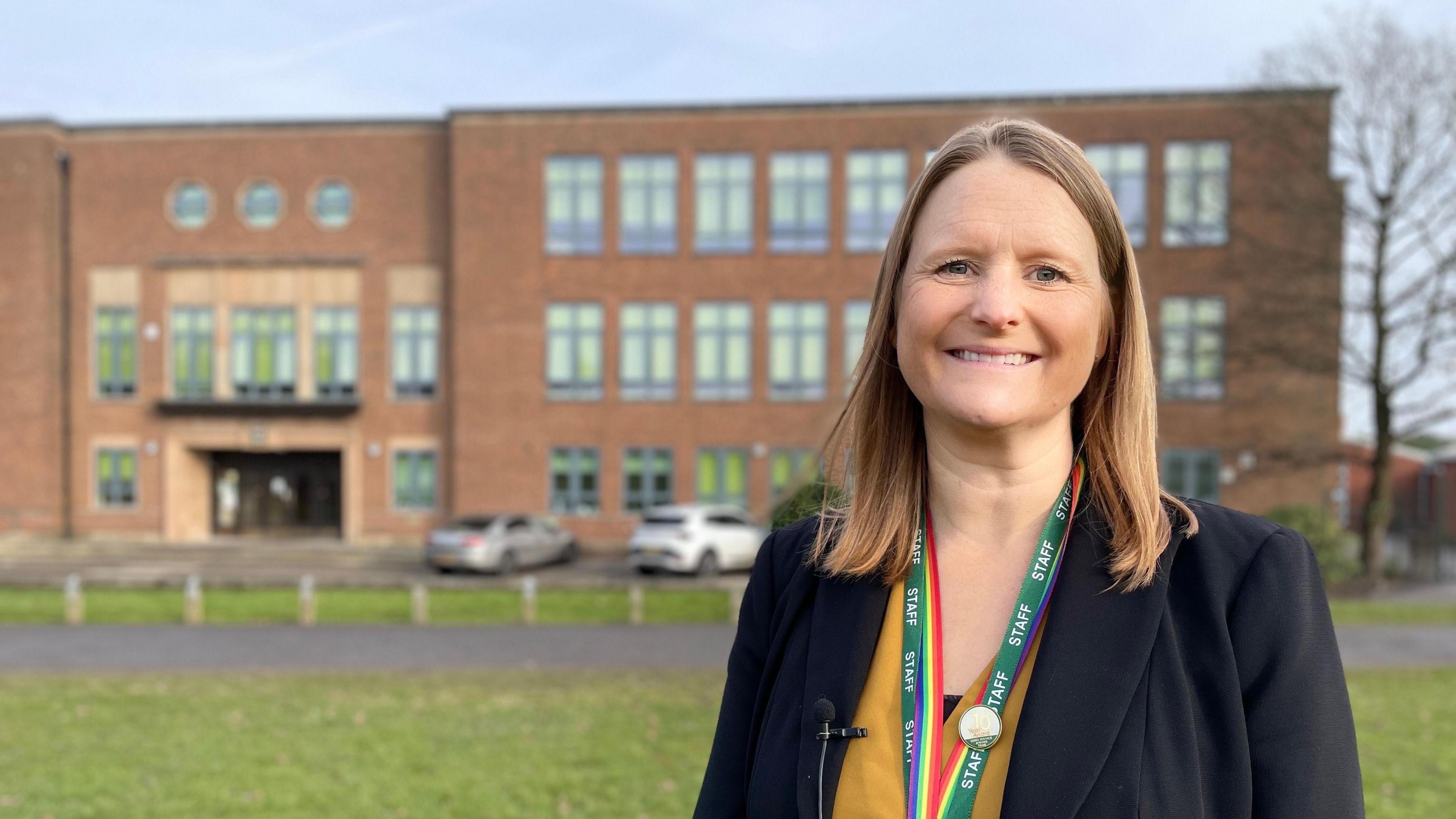 Principal Claire Smith is smiling, wearing a suit, standing in front of Tudor Grange Academy in Solihull, an imposing brick building