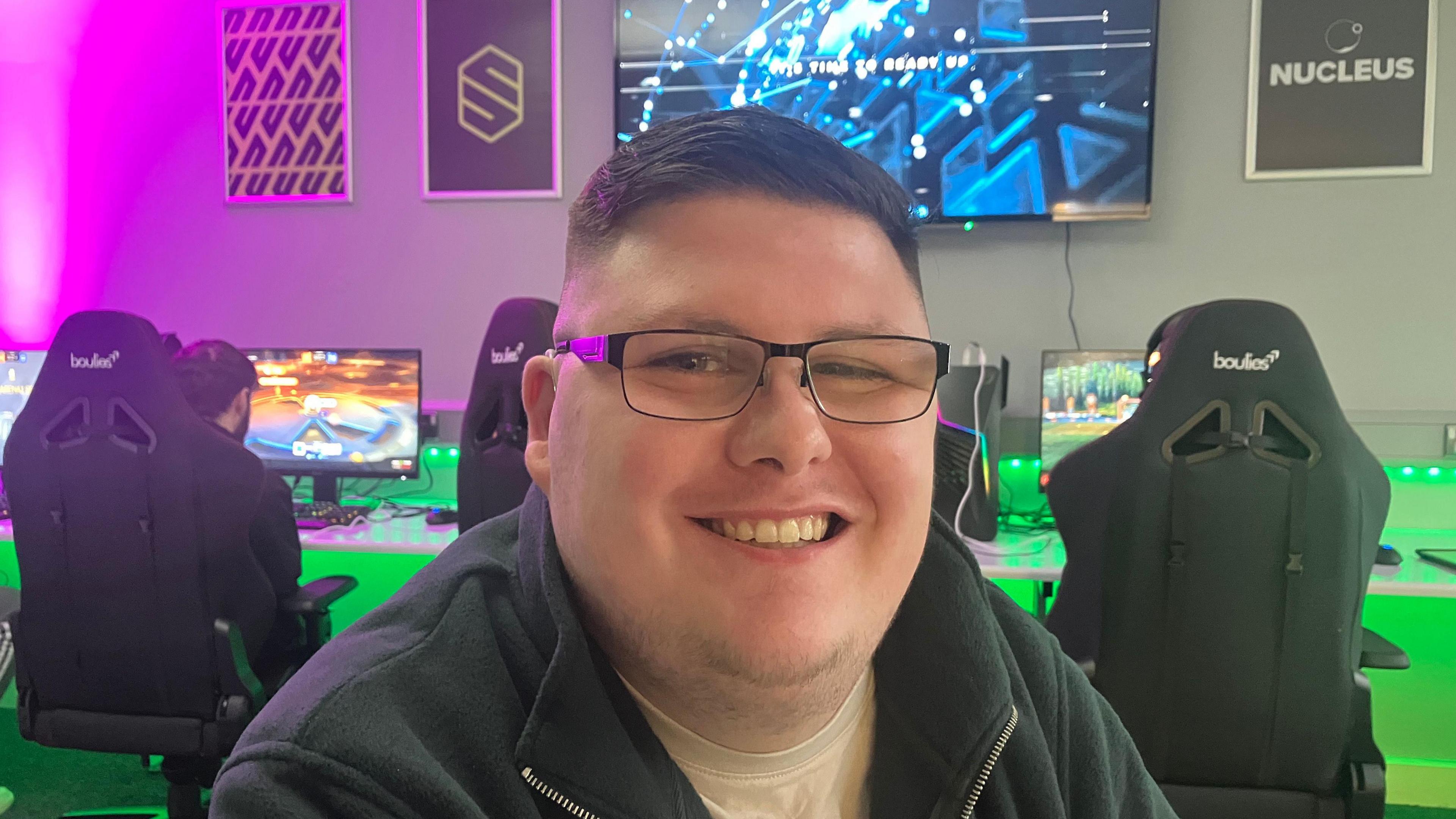 Kyle Faulkner smiles, he has black glasses. Behind him there are gaming chairs and gaming monitors. There is a large monitor on the wall.