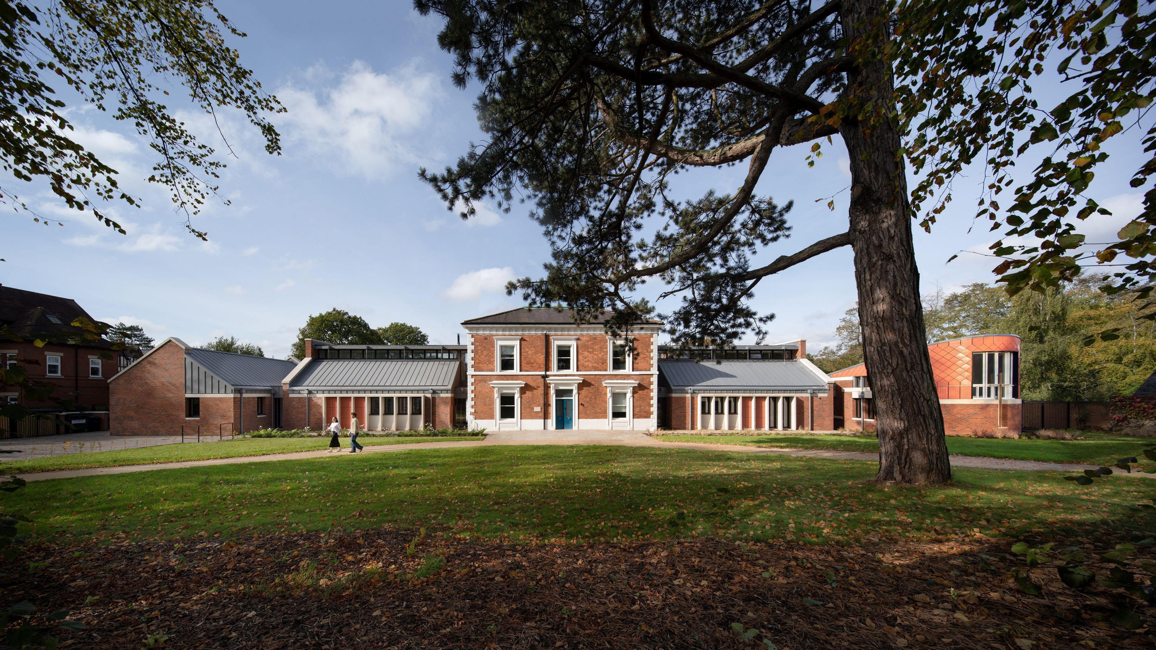 A red brick building with white windows and grey slanted roof which is surrounded by green space and large trees.