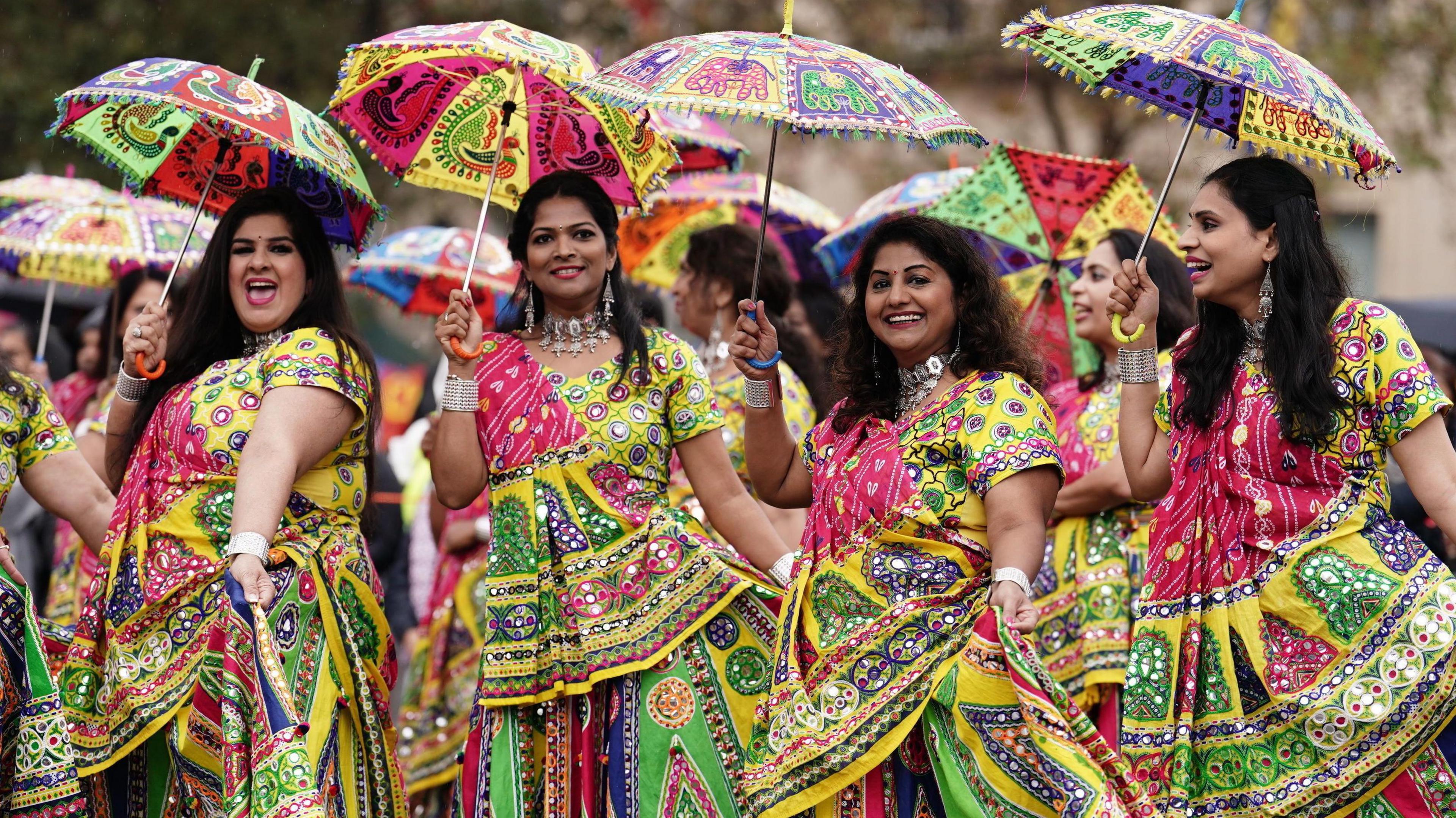 Four women laughing in colourful traditional Indian outfits holding matching colourful umbrellas. 