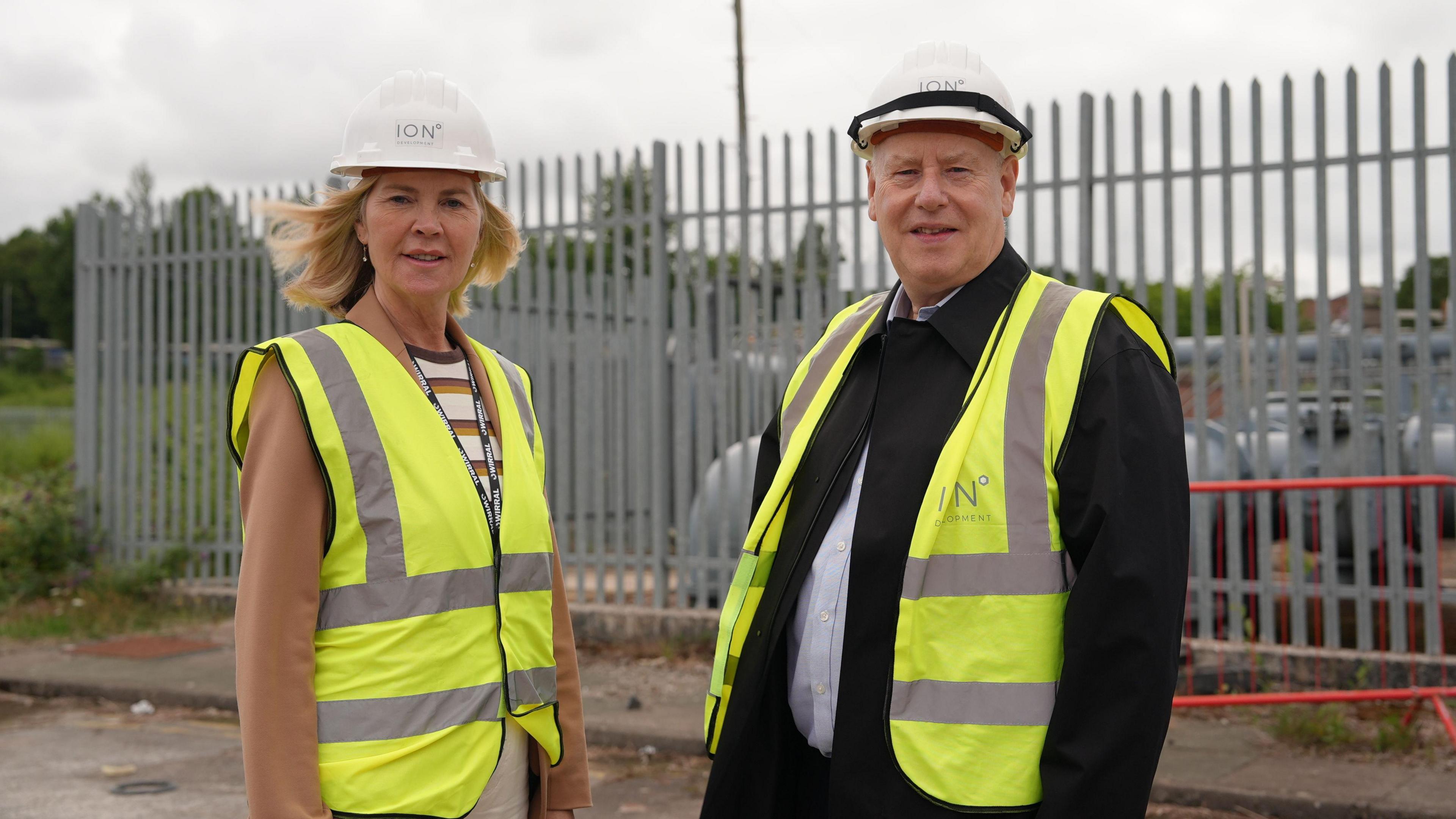 two people standing in front of metal fencing, wearing high vis waistcoats. On the left, a blonde woman in a hard hat and on the right a man wearing a suit under the high vis and a hard hat