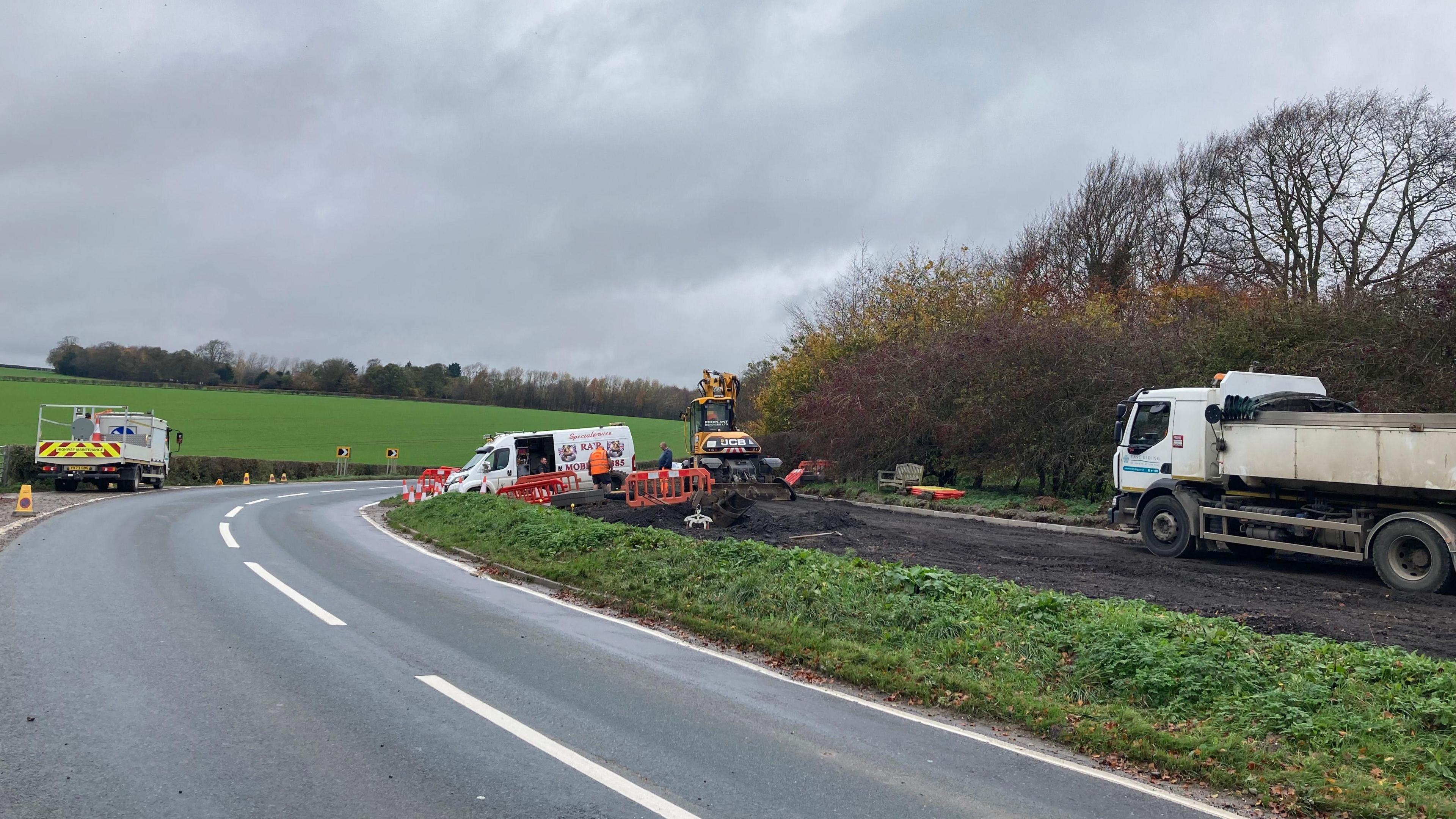 A road which has no cars driving along it bends round to the right. On the right a grass verge can be seen with maintenance vehicles and a digger which has taken up some of the grass to leave a neat line of soil. Cones can also be seen dotted around the road.
