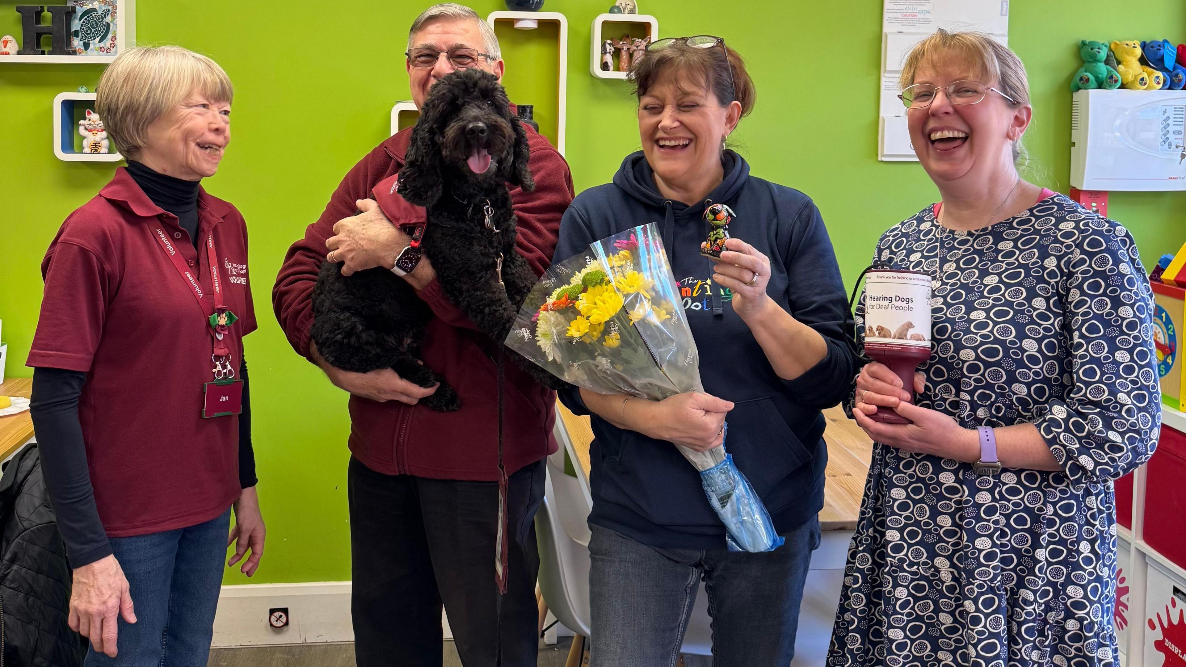 A mixed group of people stand in a bright green room. The two on the far left, Jan and Grahame, wear red polo shirts with the Hearing Dogs charity logo on. Grahame holds Beck the black cockapoo dog in his arm. Next to him, a woman wearing a blue hoody holds a bunch of flowers, and stood next to her, another woman in a blue dress patterned with white circles holds a collection tin.