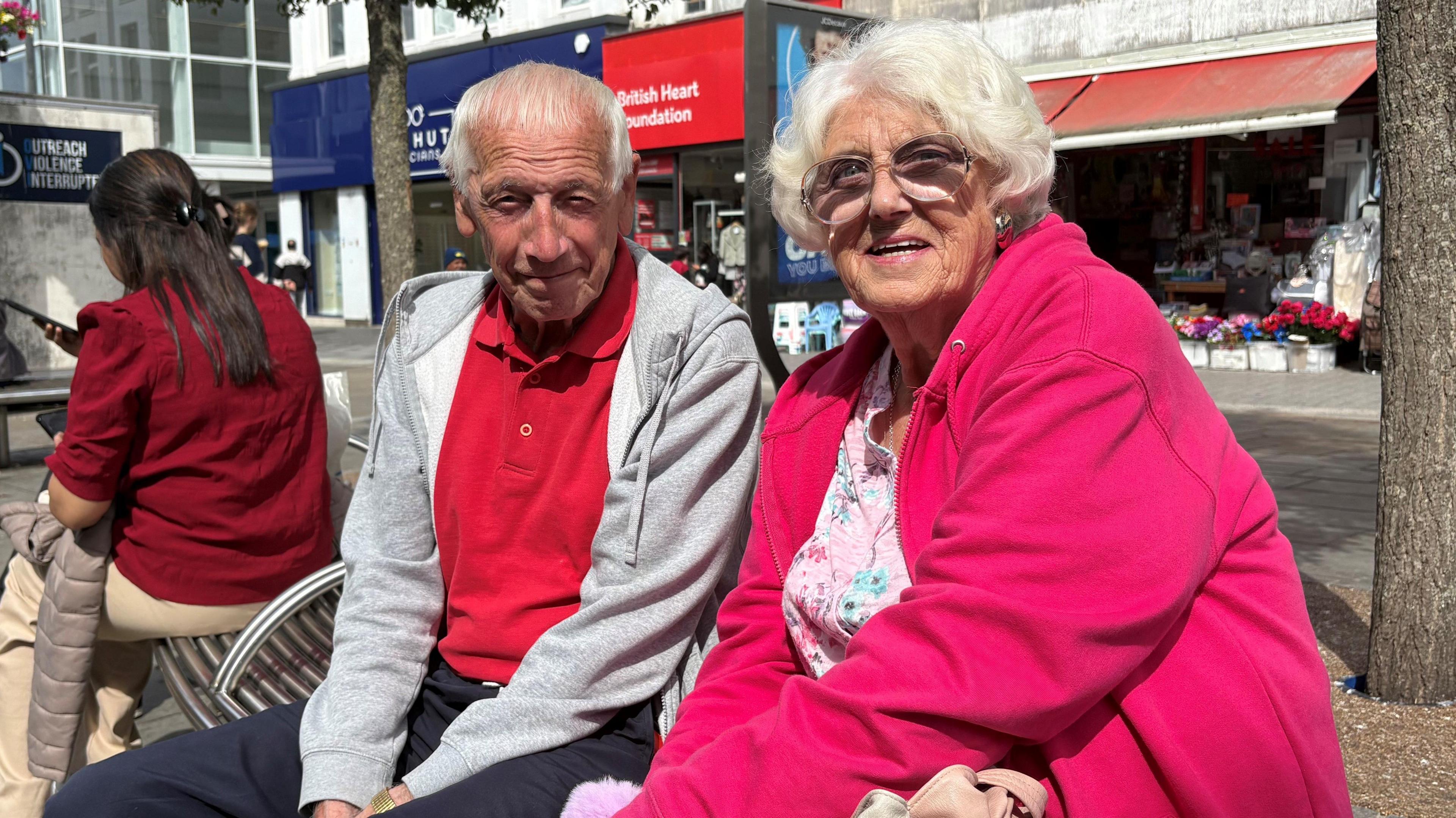 A man and woman sit on a bench in front of some shops on a sunny day.