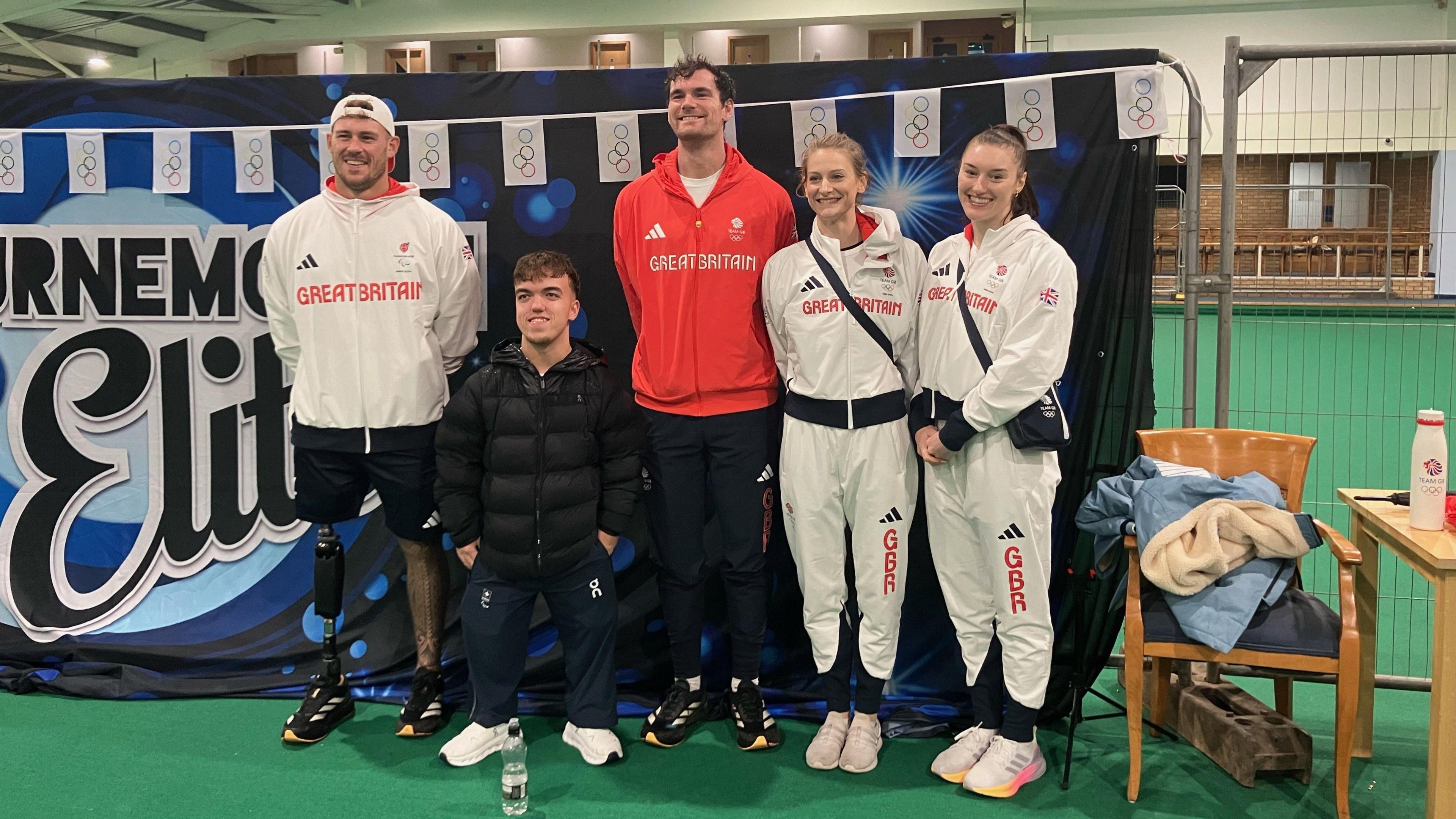 Three male athletes and two female athletes wearing 'Great Britain' sports kids smiling at the camera.