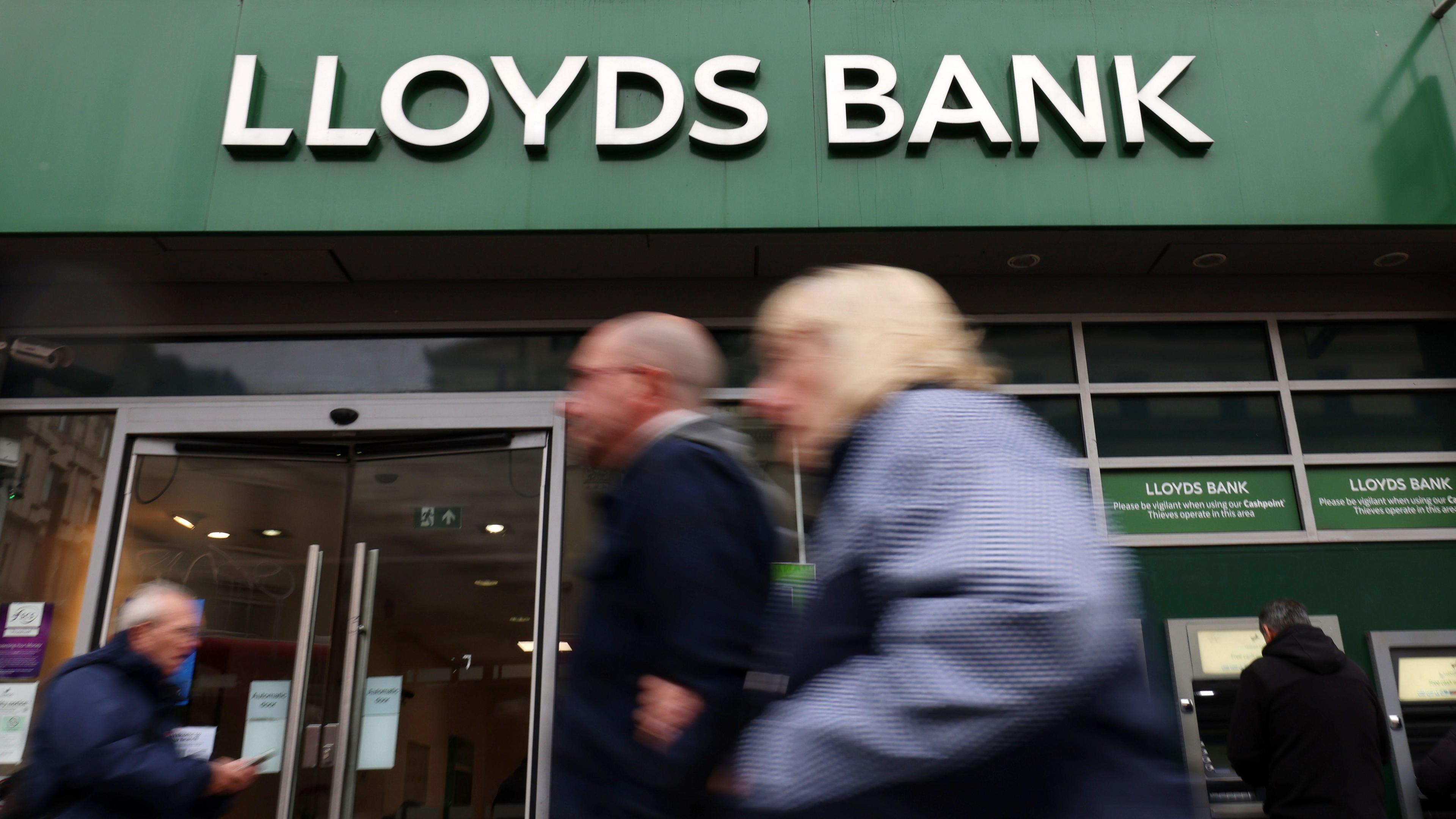 A shot of the front of a Lloyds Bank - it's branded name is in white lettering above the bank on its distinctive green background - in the foreground are a number of people walking and are out of focus, which draws focus to the bank's name - a person can also be seen taking money out of a cash machine 