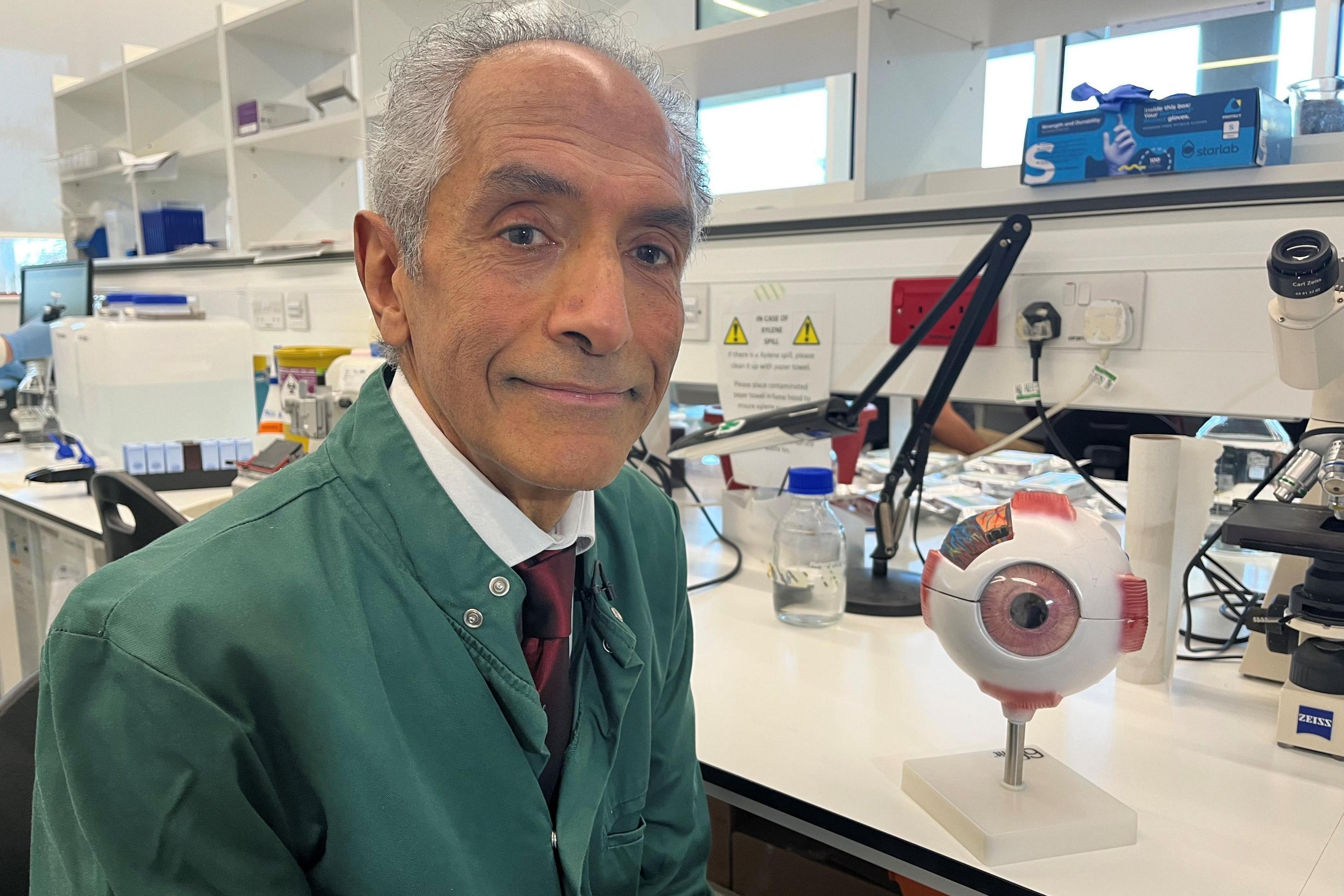 Prof Baljean Dhillon is sitting at a bench with white shelves above. He has balding grey hair and is wearing a white shirt, maroon tie and green jacket. Next to him on the bench is a model of an eyeball and a microscope, among other items