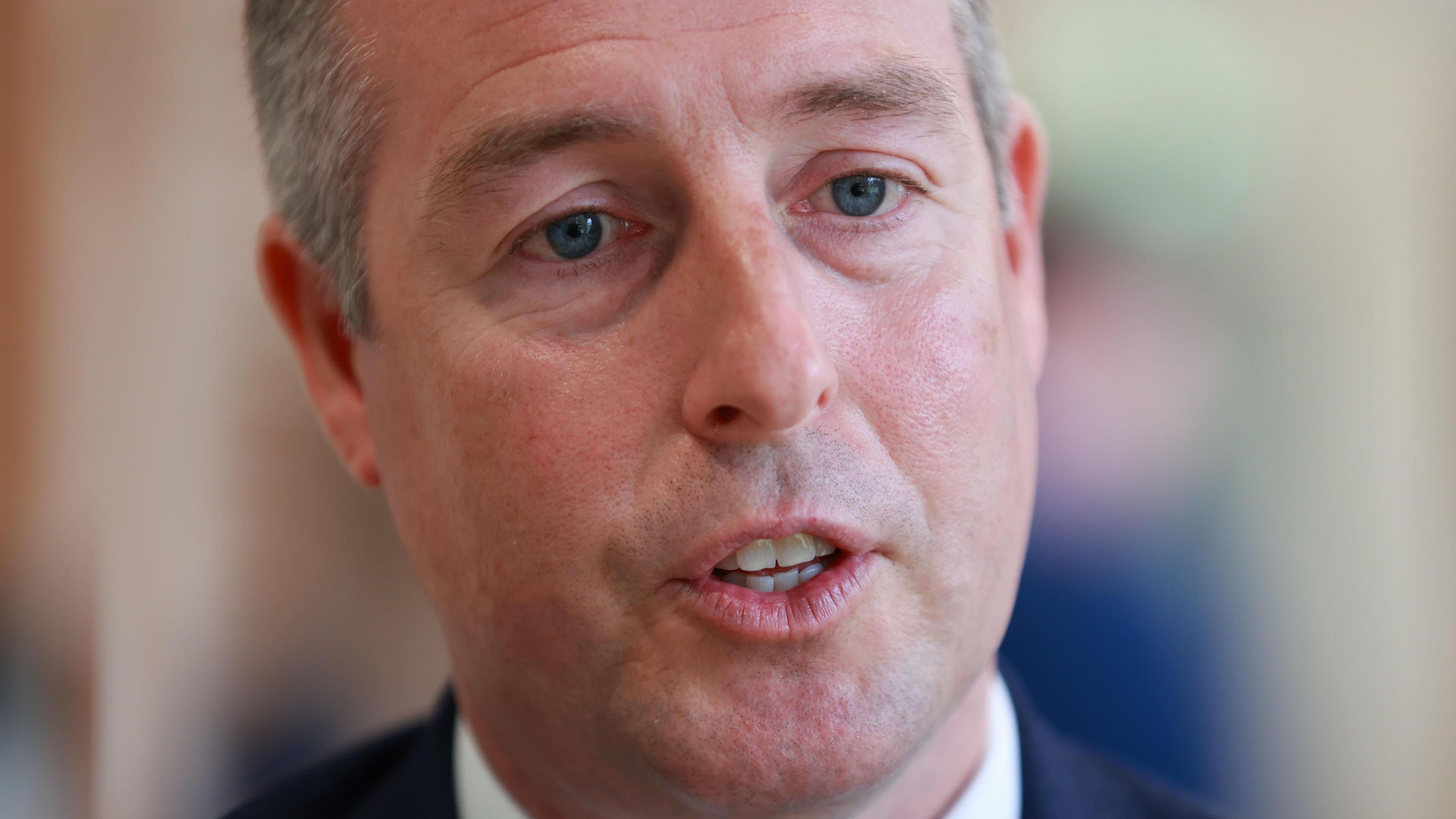 A close up photo of Paul Givan while he is speaking.   He has short, greying hair and is wearing a navy suit and a white shirt.