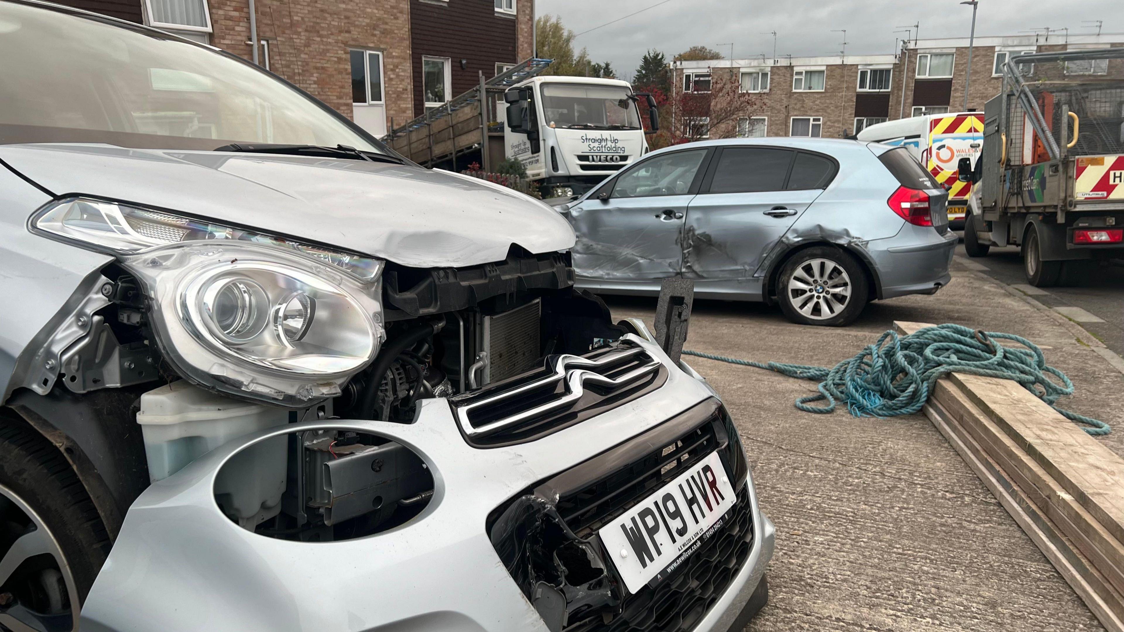 Two cars are pictures badly damaged with one having its front bumper severaly broken and another with deep scrapes down the side. The lorry involved is visible in the background