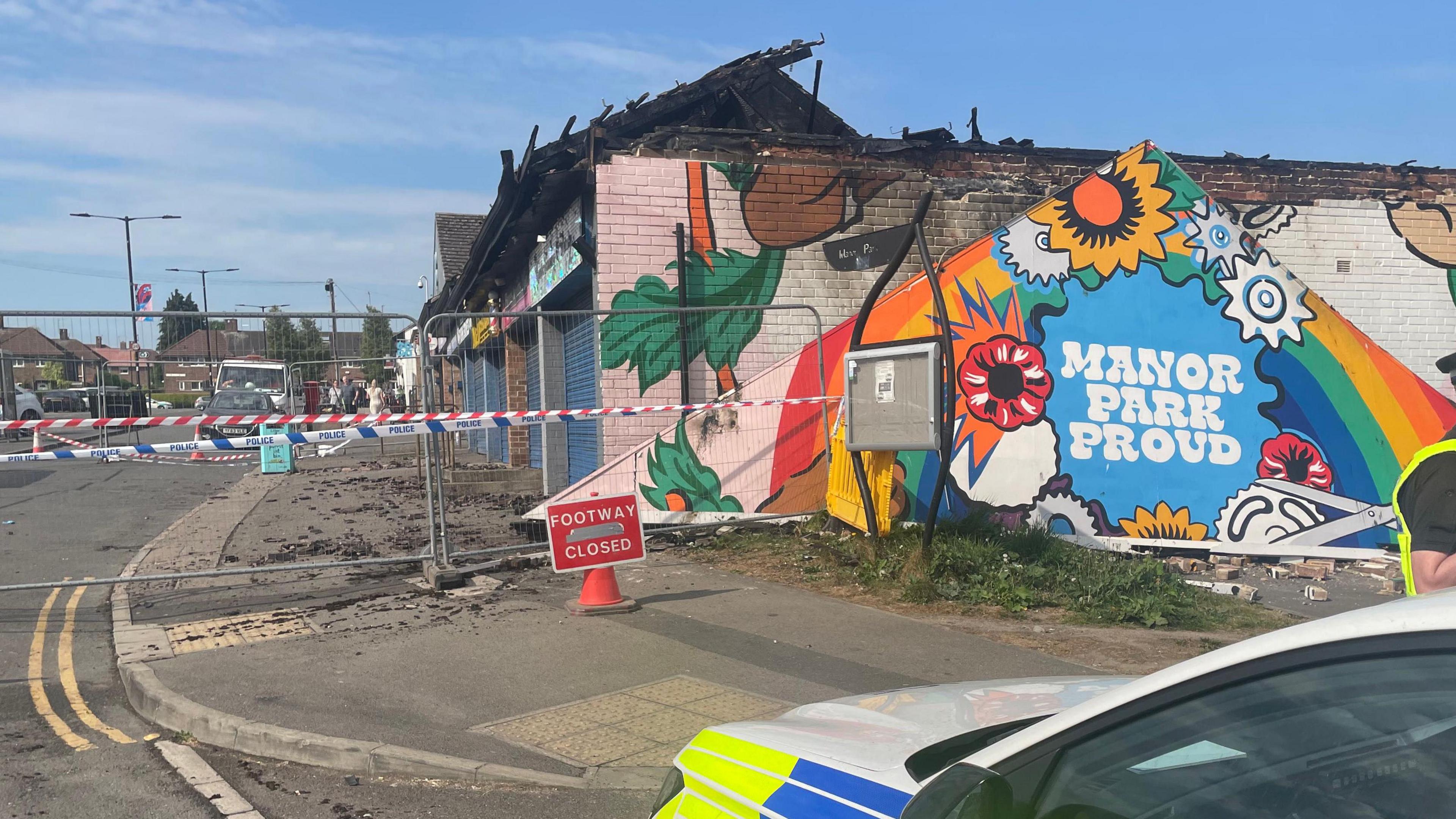 Tiles and bricks from the affected buildings litter the pavement at Manor Park Centre, off Harborough Road. Part of a mural is visible on the left hand side. The shops all have blue shutters down. A fence is up with police tape across it.