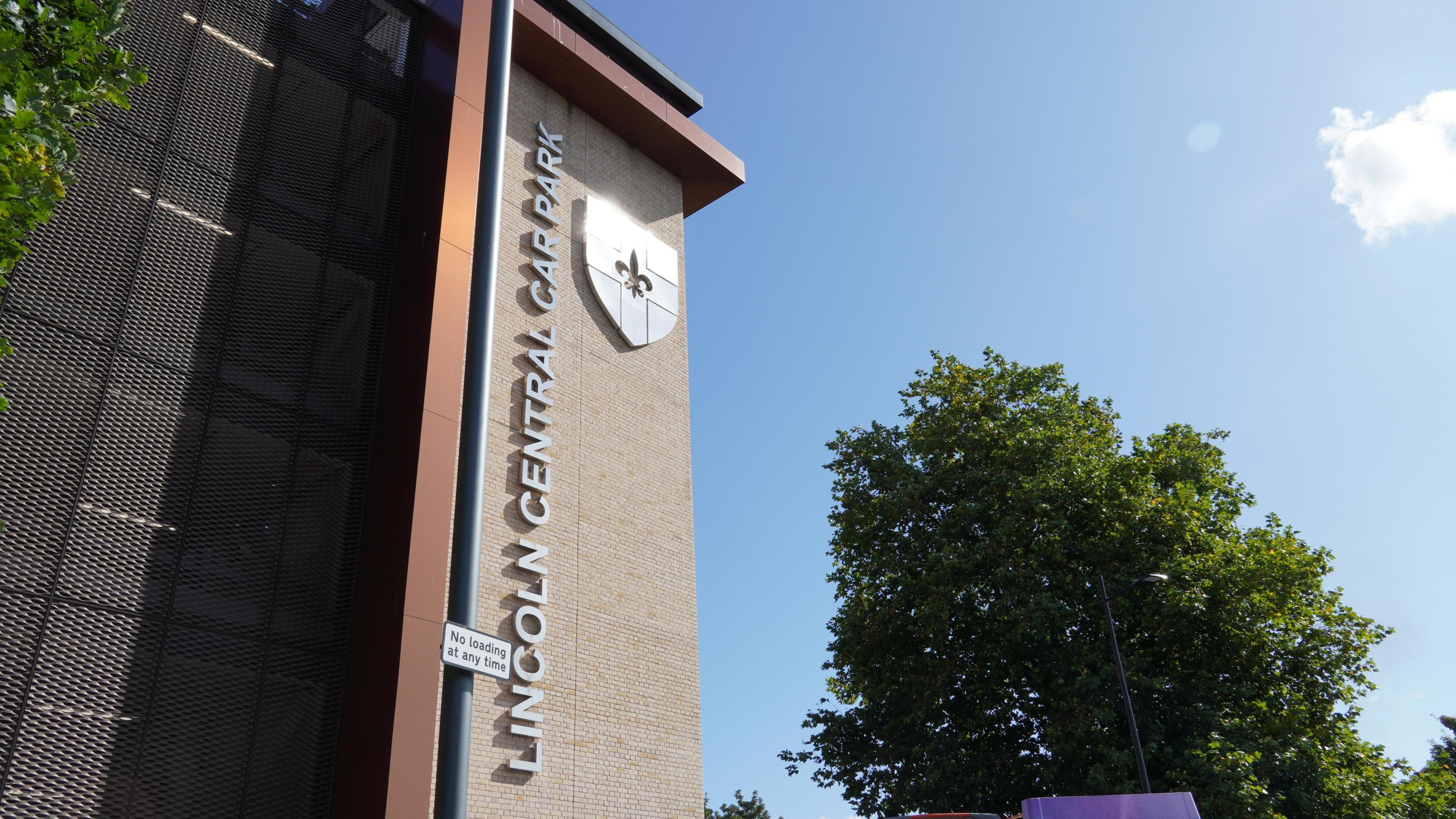 The outside of Lincoln Central Car Park, a tall, modern-looking multi-storey building, of light-coloured bricks and metallic cladding, with "Lincoln Central Car Park" advertised in large silver letters alongside a silver City of Lincoln crest.