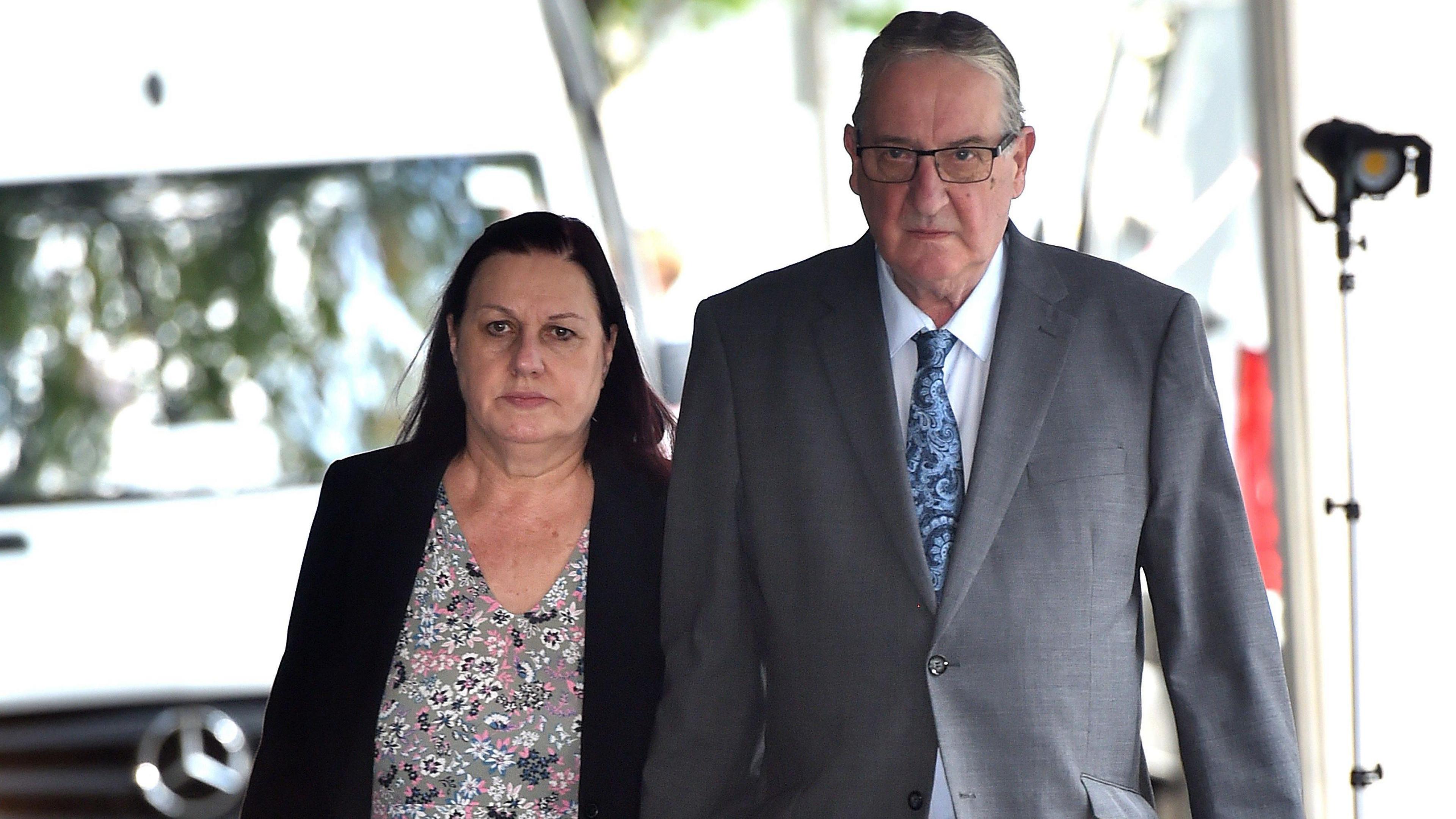 Susan Letby, who has dyed red hair and a wears a dark blazer over a flowery blouse, and John Letby, who has short grey hair and wears a light grey suit, walk towards Manchester Crown Court hand in hand with stern expressions on their faces 