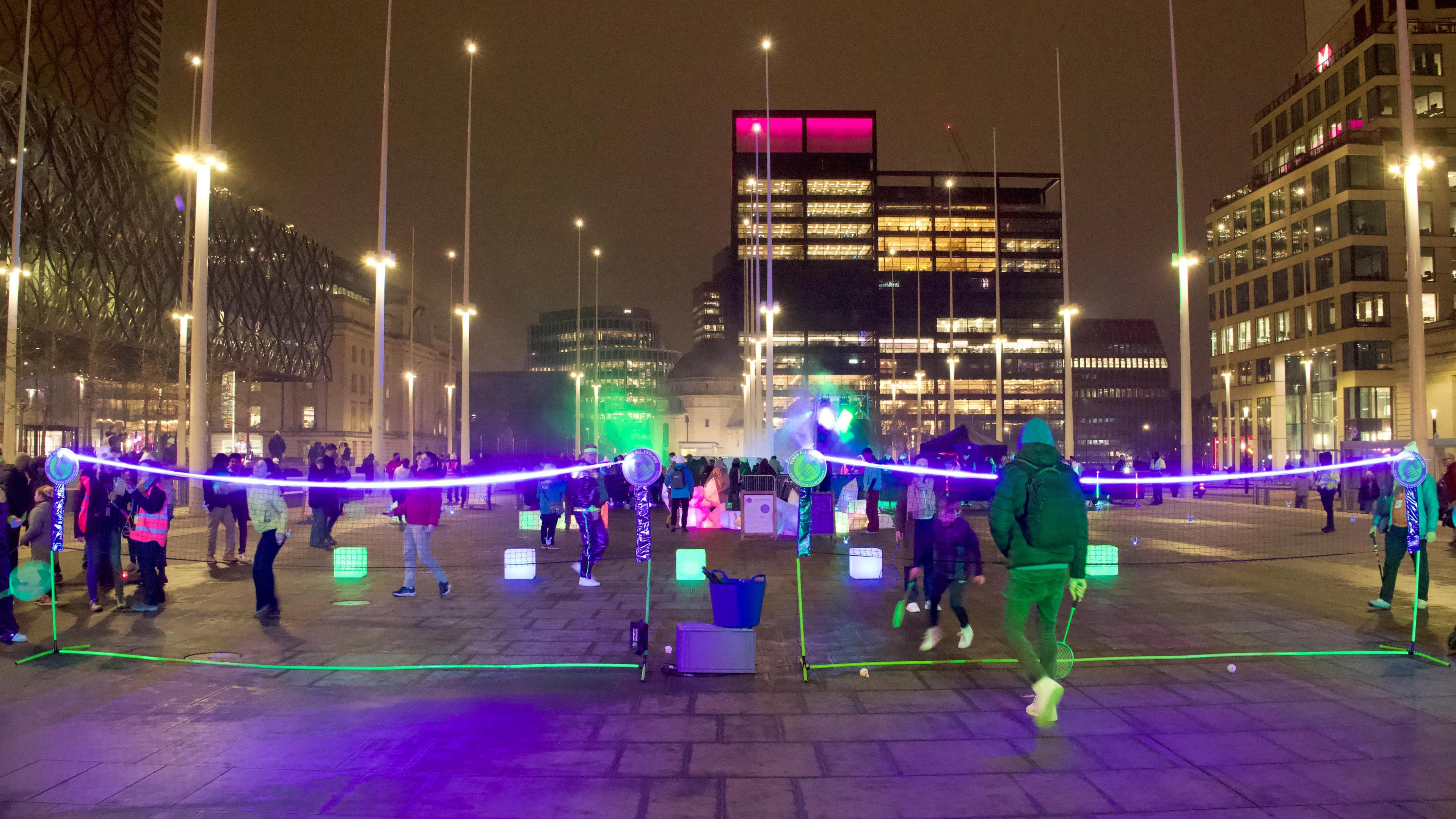 People playing on two badminton courts in a square. The net is illuminated and so are the shuttlecocks