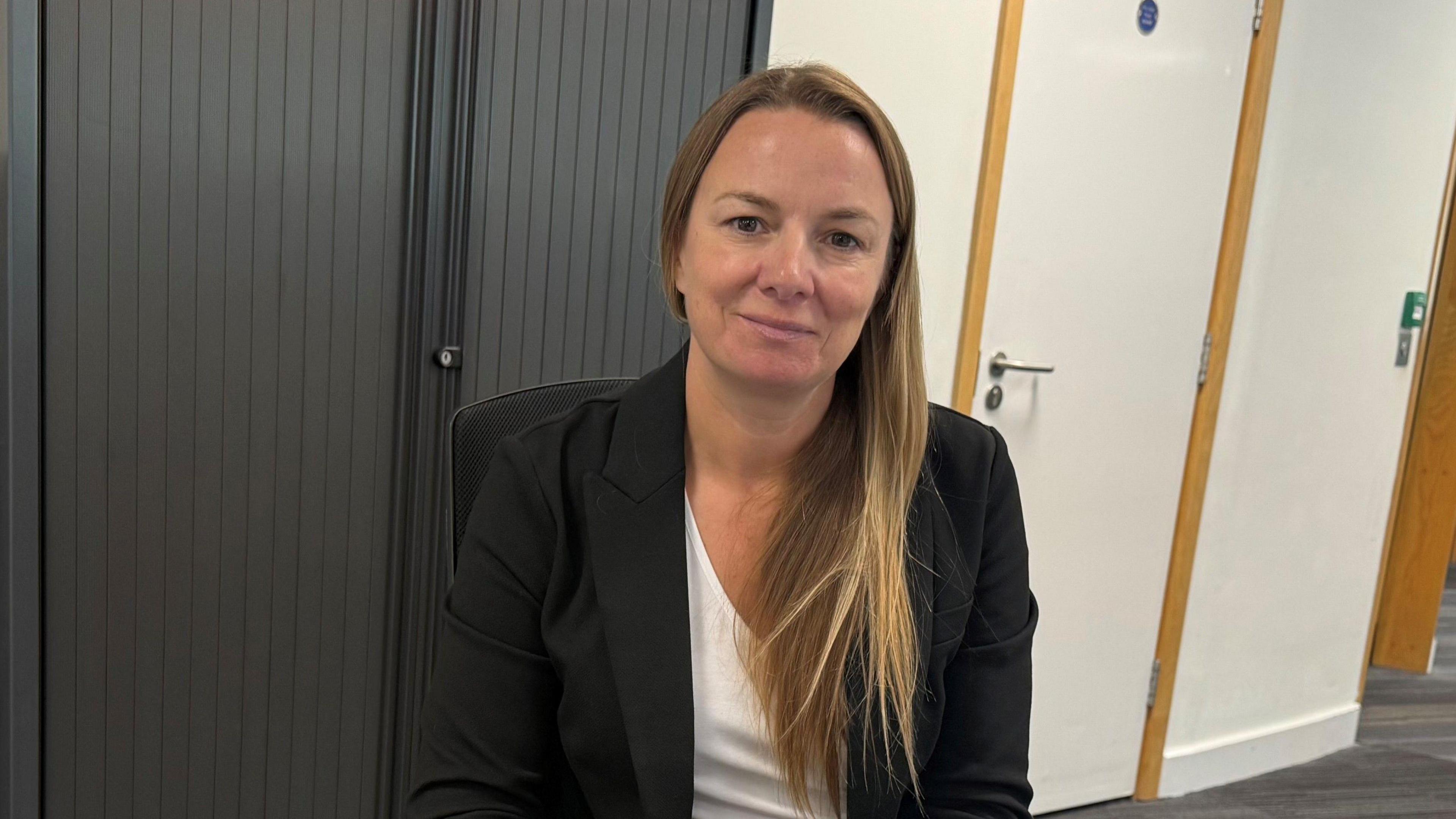 Gillian has long blonde hair, dressed in a black blazer over a white top, sits in an office setting. Behind her is dark vertical blinds and a white door with a silver handle. The carpeted floor and a glimpse into another room through an open doorway are visible on the right.