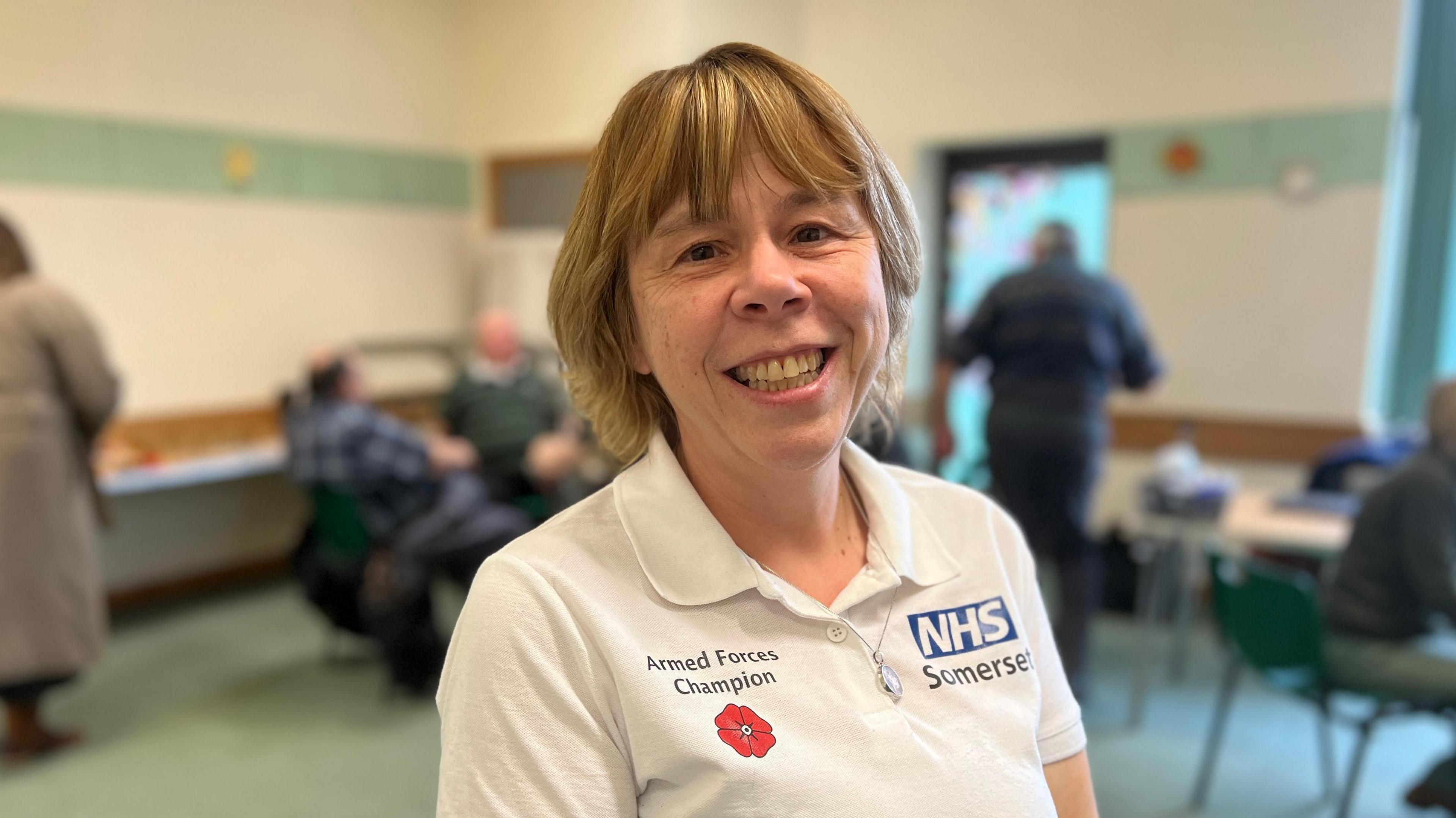 Blonde-haired women smiling at the camera. She has a white T-Shirt on saying Armed Forces Champion and a big NHS Somerset logo on it.