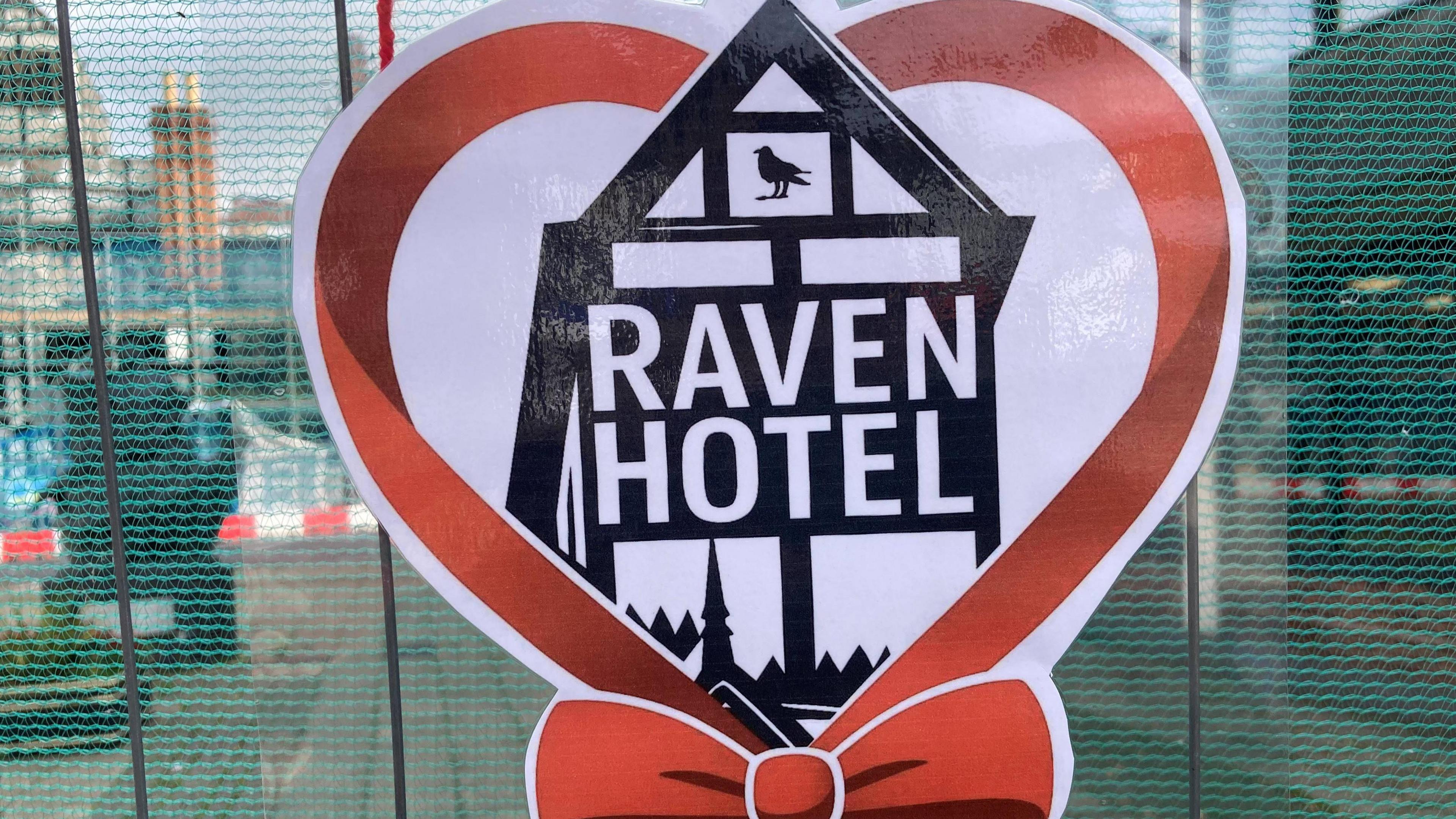 A large logo of a red ribbon, with an artist's illustration of the Raven hotel in the middle - the logo has been attached to steel fencing in front of a derelict building.