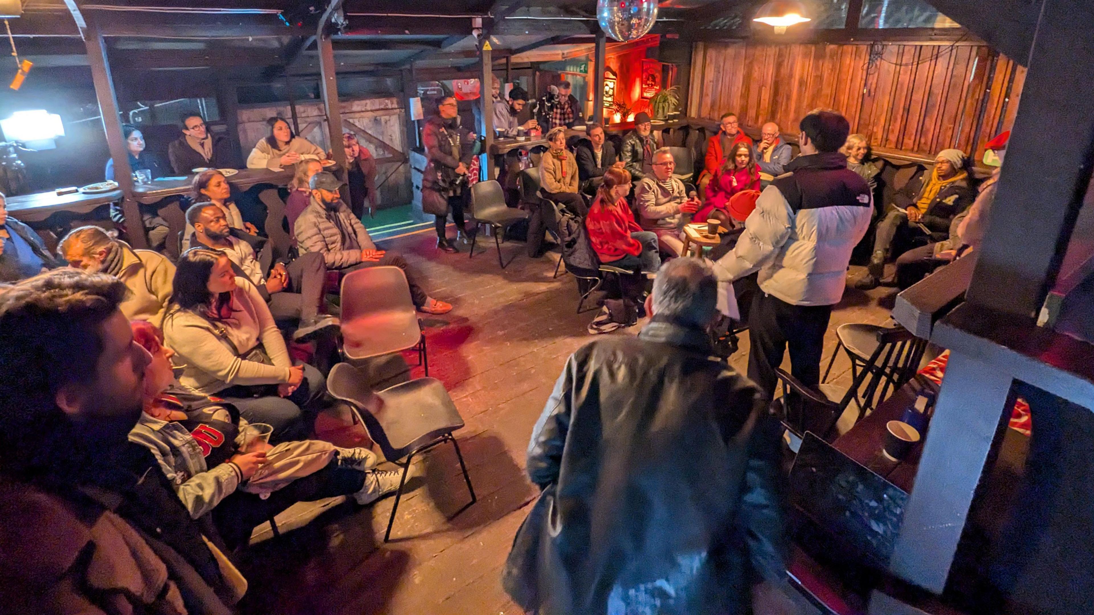 A group of people in a room listening to a man talking, who has his back to the camera. The man is wearing a cream and black cat, and has short dark hair. The group of people are in a room with lots of chairs and most are sitting down. 