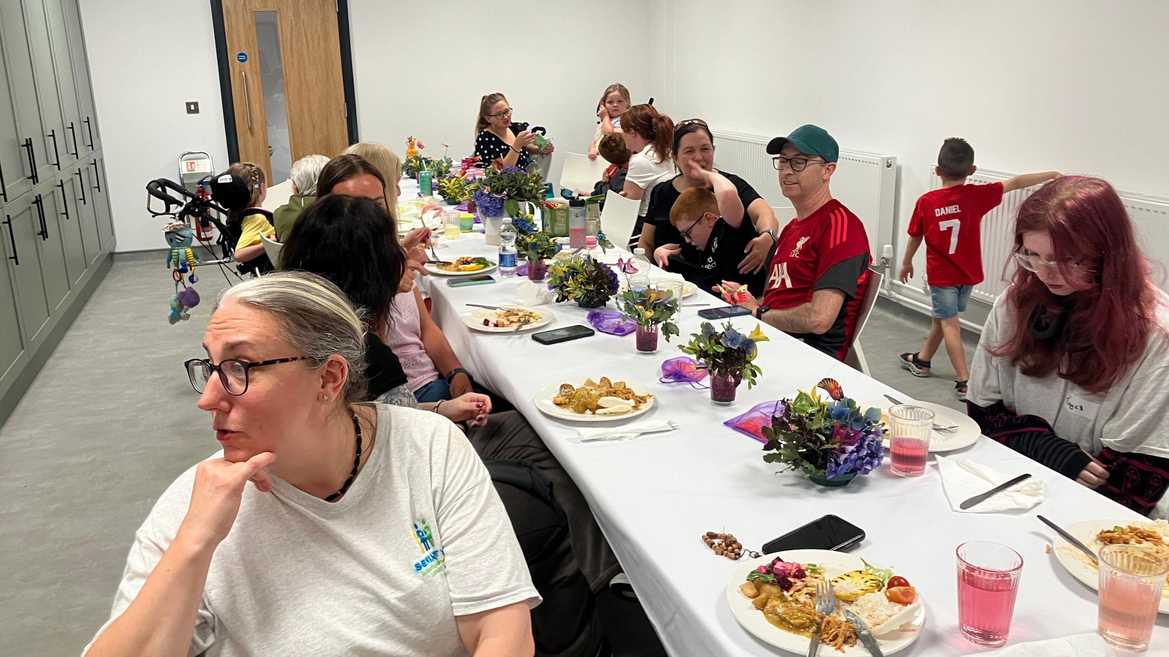 A long white table sits a dozen people. There are plates of food in-front of each person. The table is in a small white-walled room. There are half a dozen bunches of flowers down the centre of the table.  