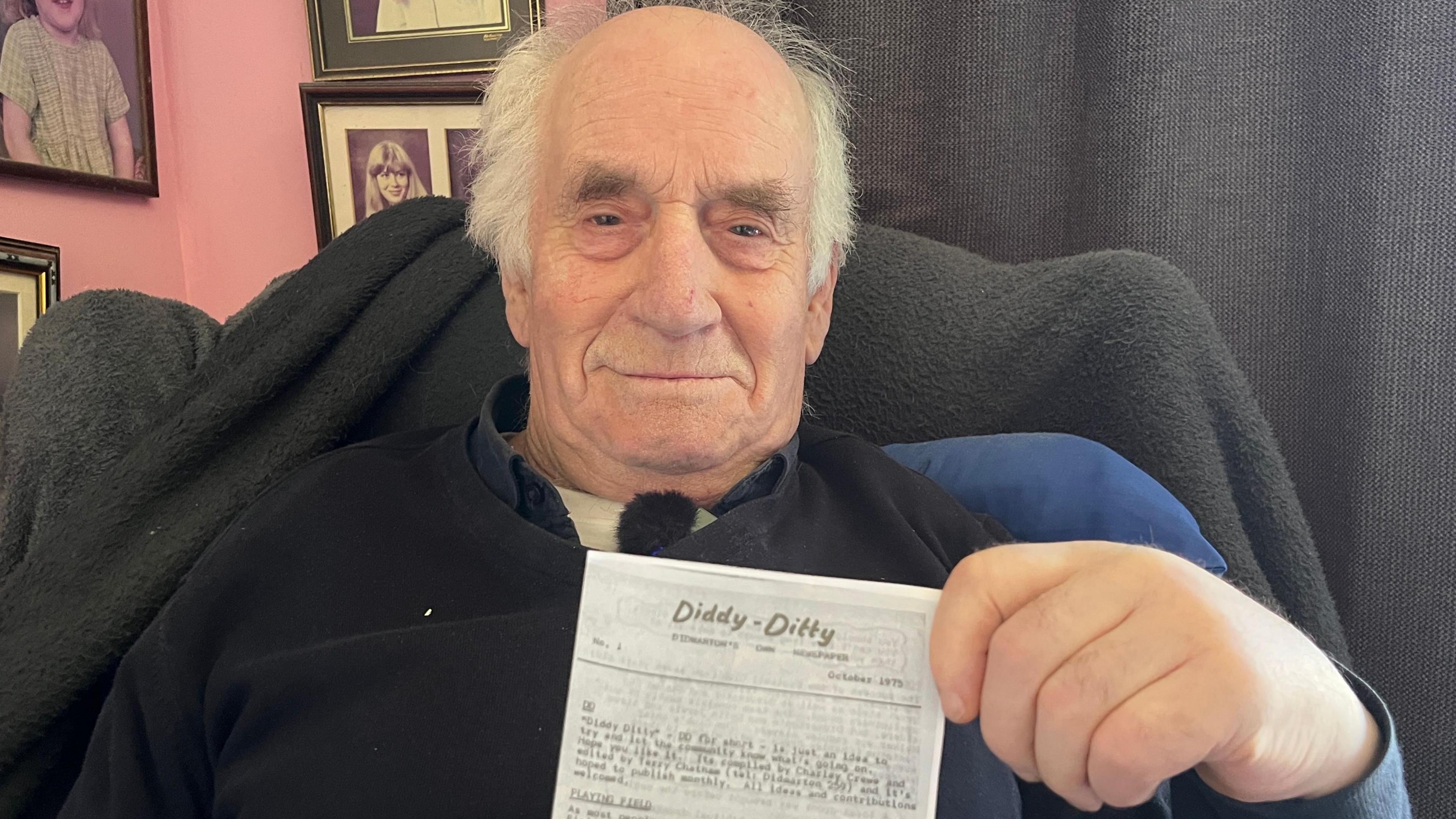 Man with grey hair smiles whilst holding up a copy of the first edition of a village newspaper called Dyddi - Ditty