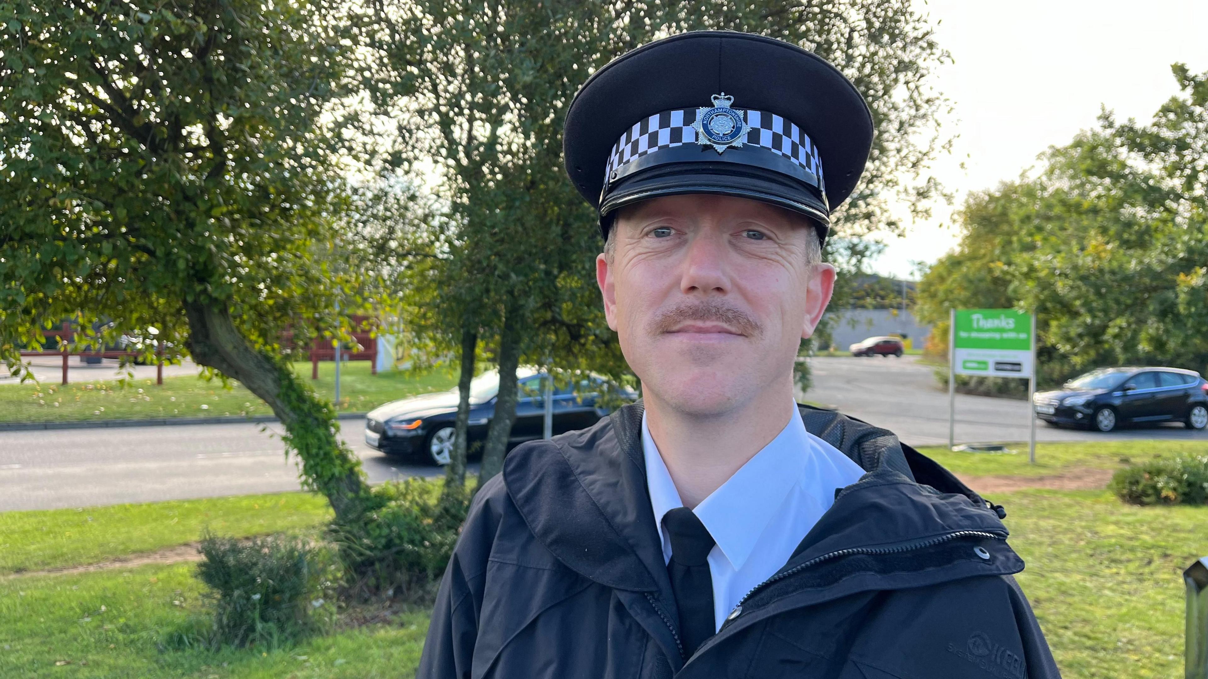 Male police officer with black coat and police hat on, standing in front of trees, with a green grass background.