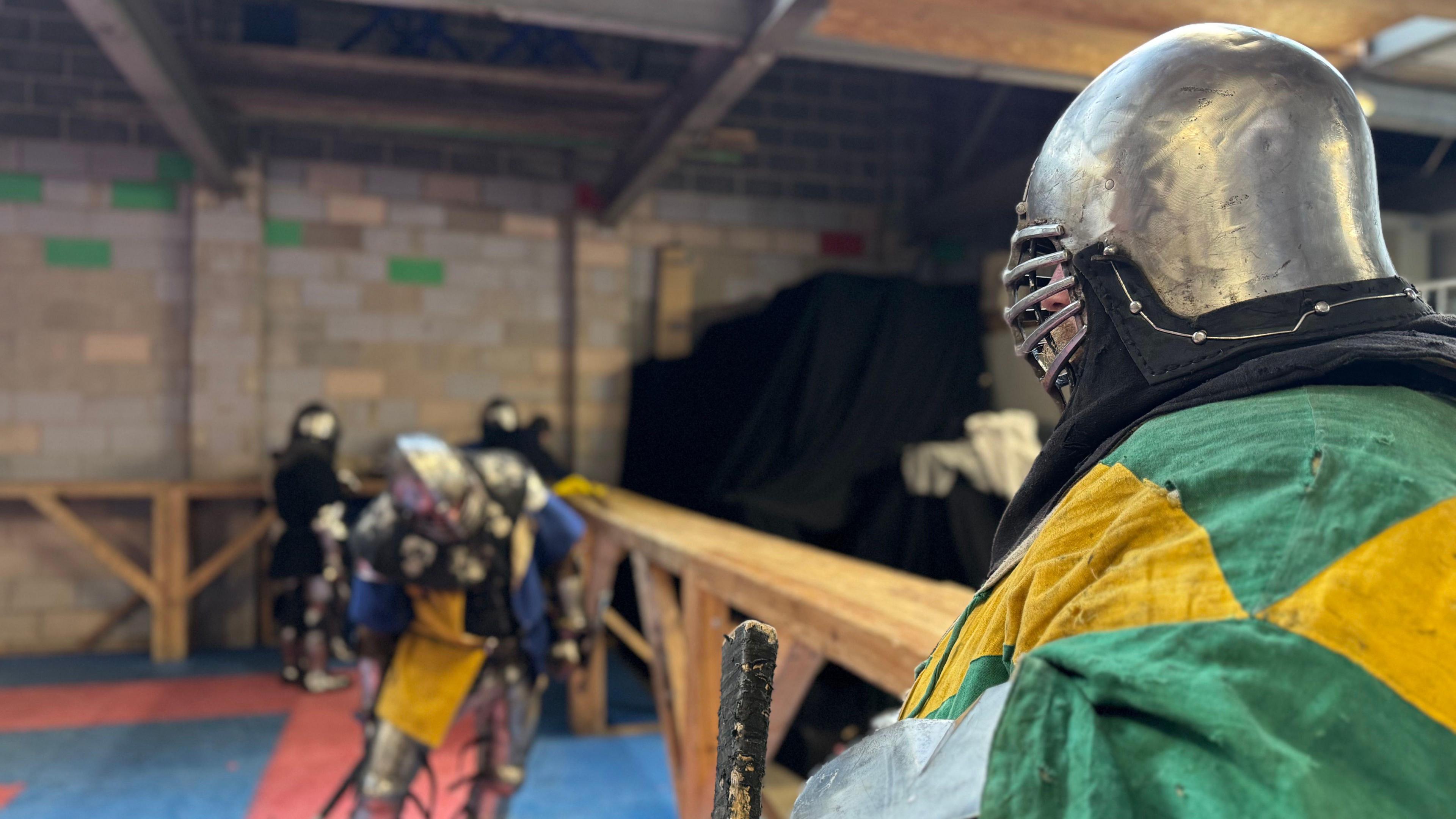 A man in a helmet and a colourful tunic looks into the distance. He's leaning on a wooden barrier with other armoured men in the background.