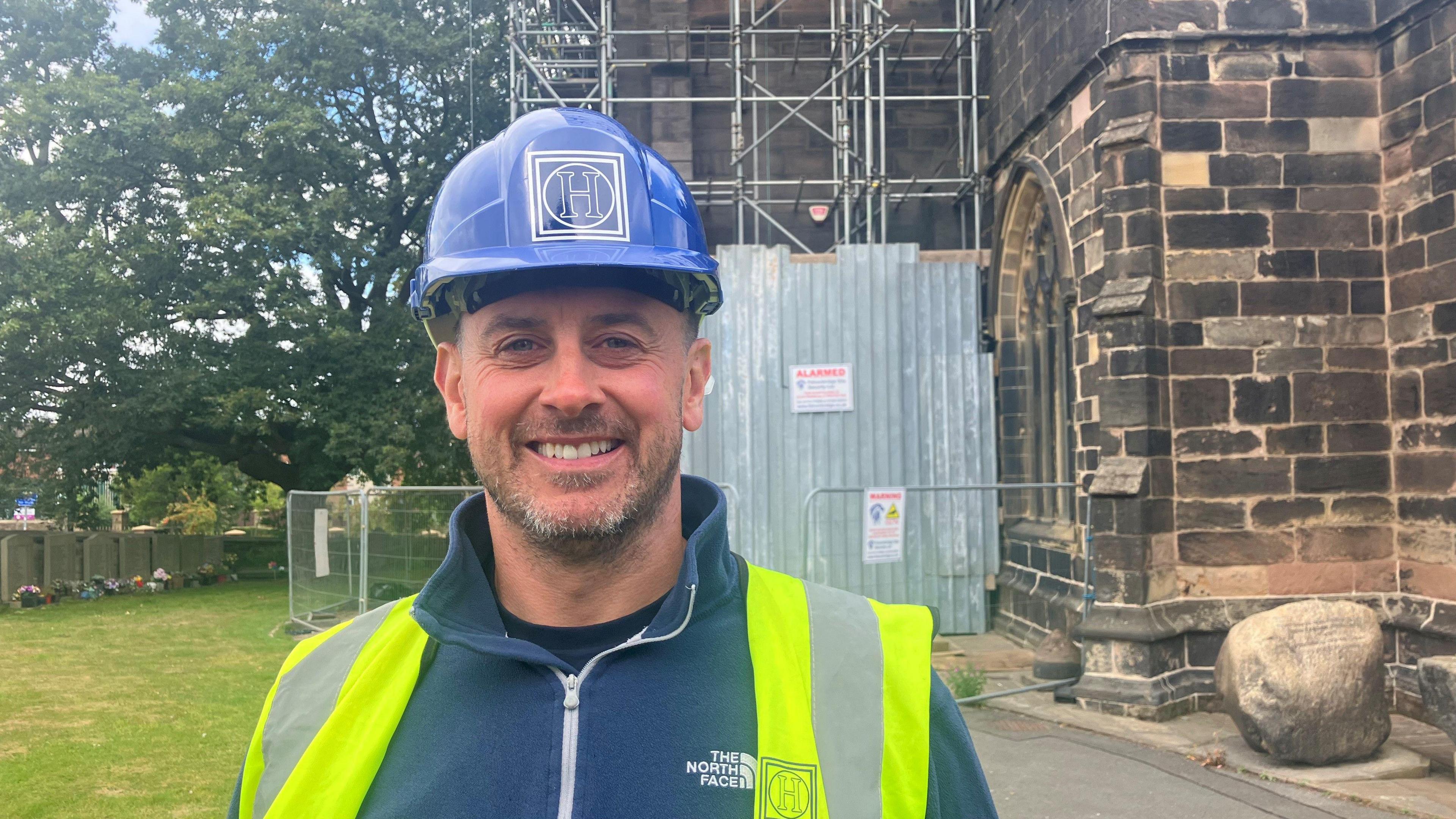 Photograph of James Dalton from Heritage Conservation Restoration Ltd. He is pictured in the church yard of St Wilfrid's in Standish. He is wearing a blue hard hat and yellow-green high visibility jacket.