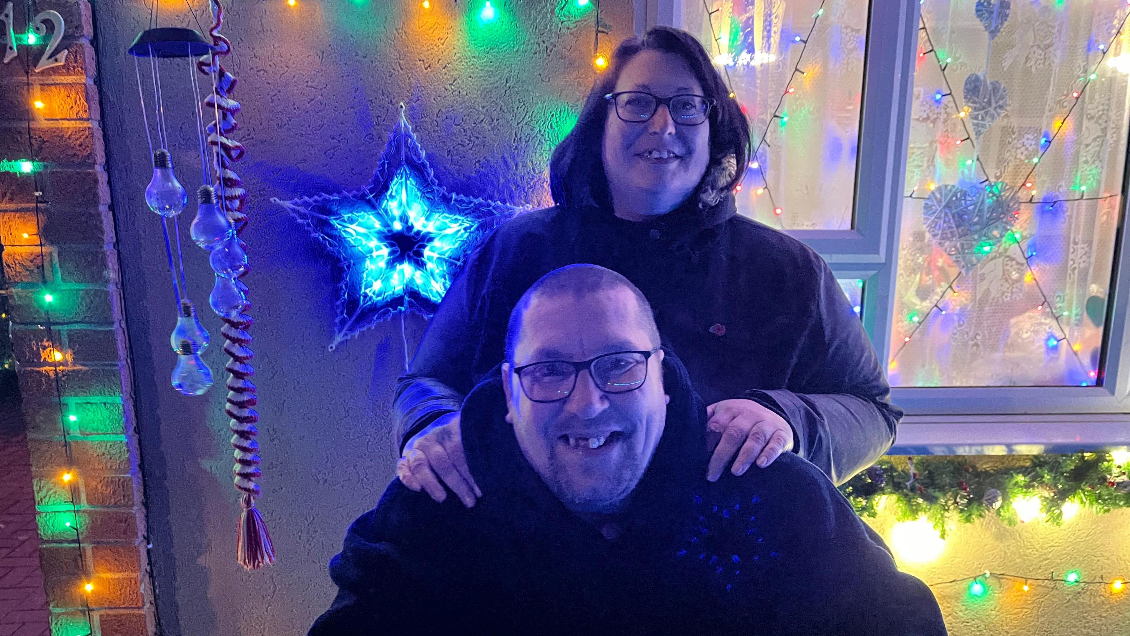 Kerri Frost in a black coat with her hands on Mark's shoulders. He is wearing glasses and a black hoodie. They both stand in front of a front door that is lit up with lots of different coloured lights. 