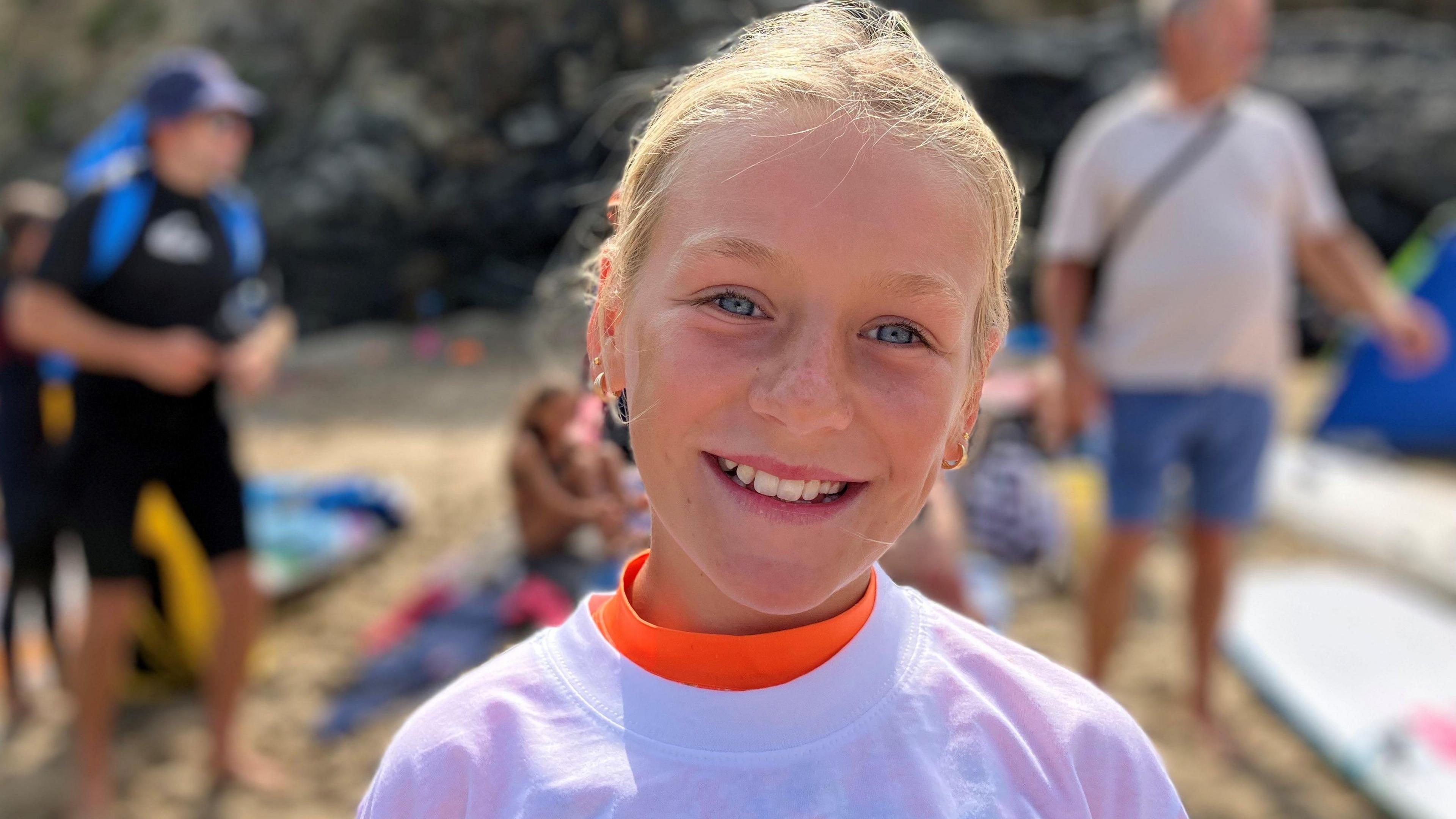 Young girl with blue eyes and blonde hair tied back, smiling at the camera on the beach