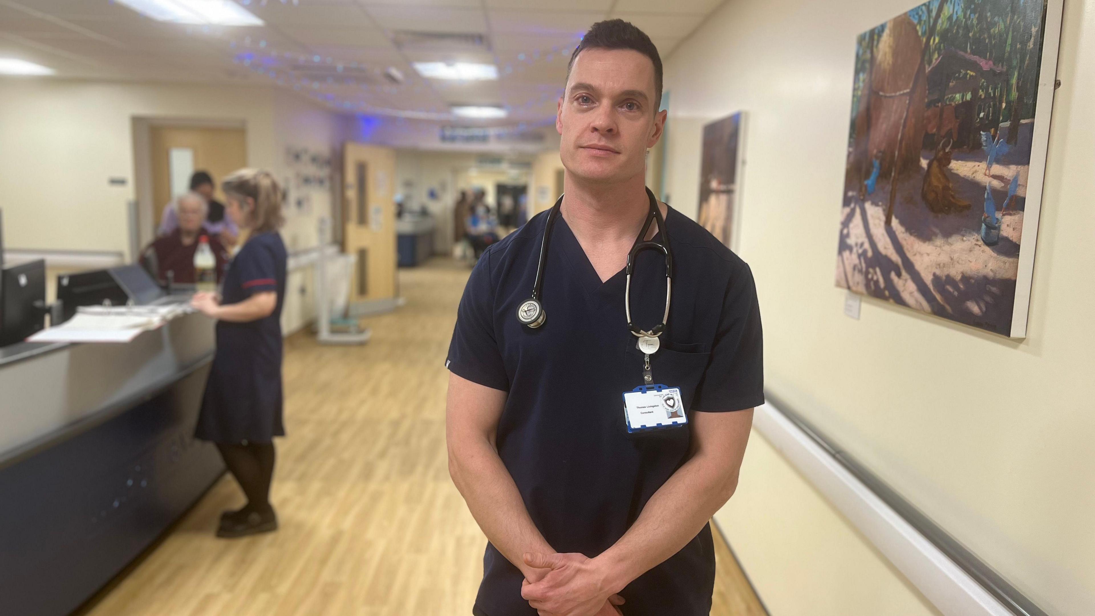 Man in scrubs stands in hospital ward corridor 