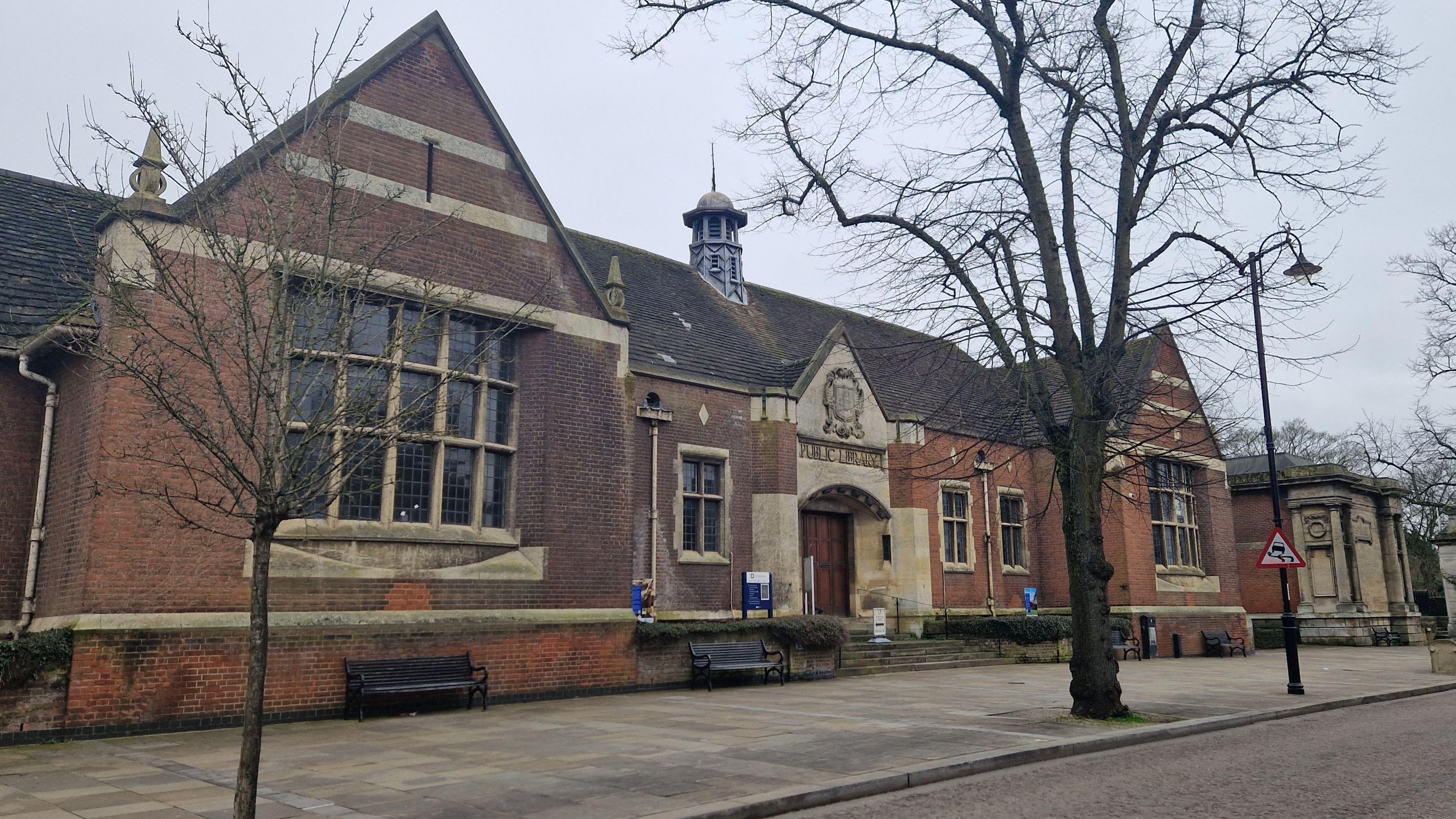 Single-storey red brick and stone building - with small windows framed by stone and a stone entrance area. There is a coat of arms over the door and the words "public library" are just visible. There are paving slabs and trees in front of the building.