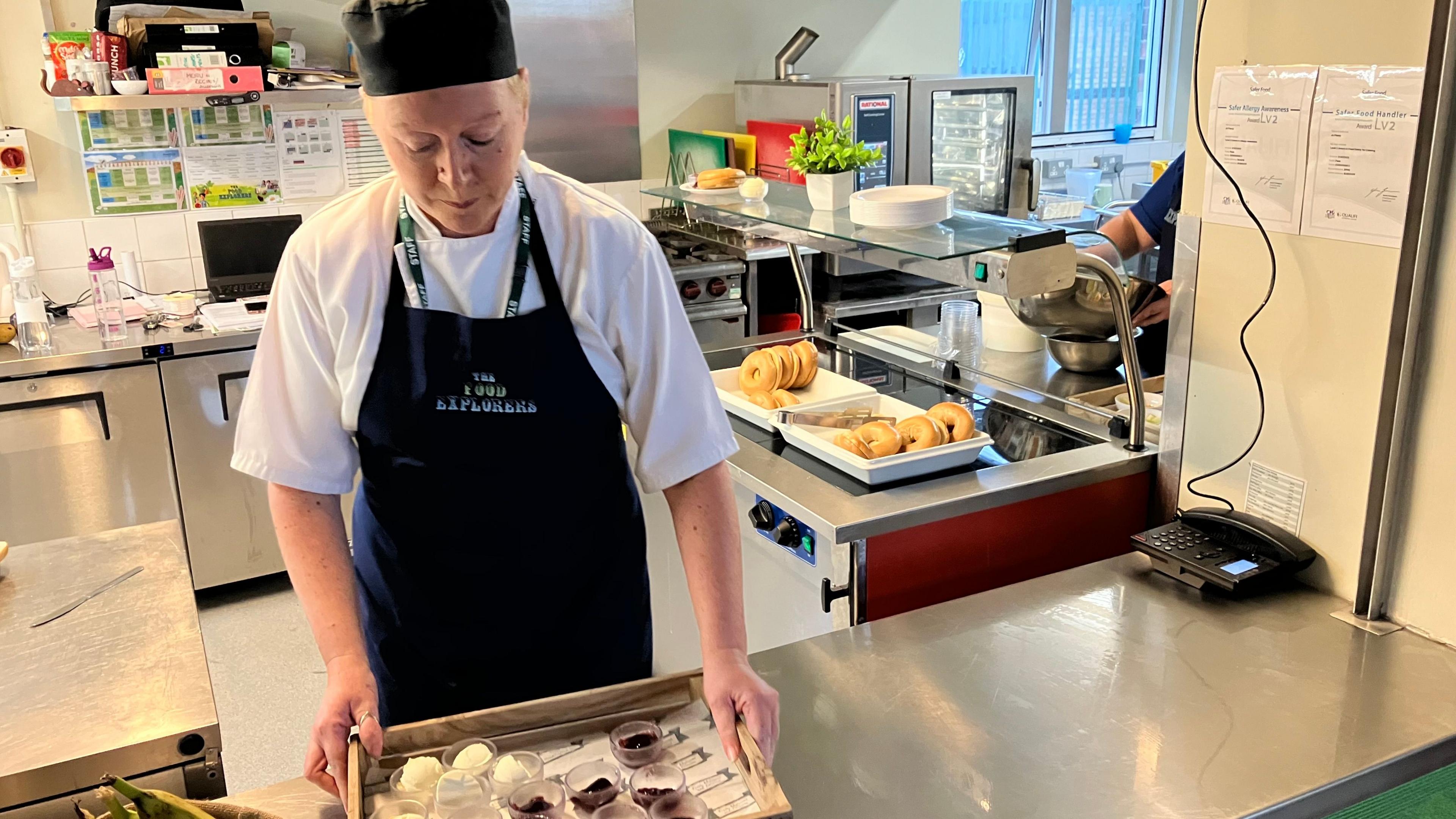 A caterer wearing a black apron stands in a school kitchen, holding a tray of what appears to be jelly in small glass dishes.