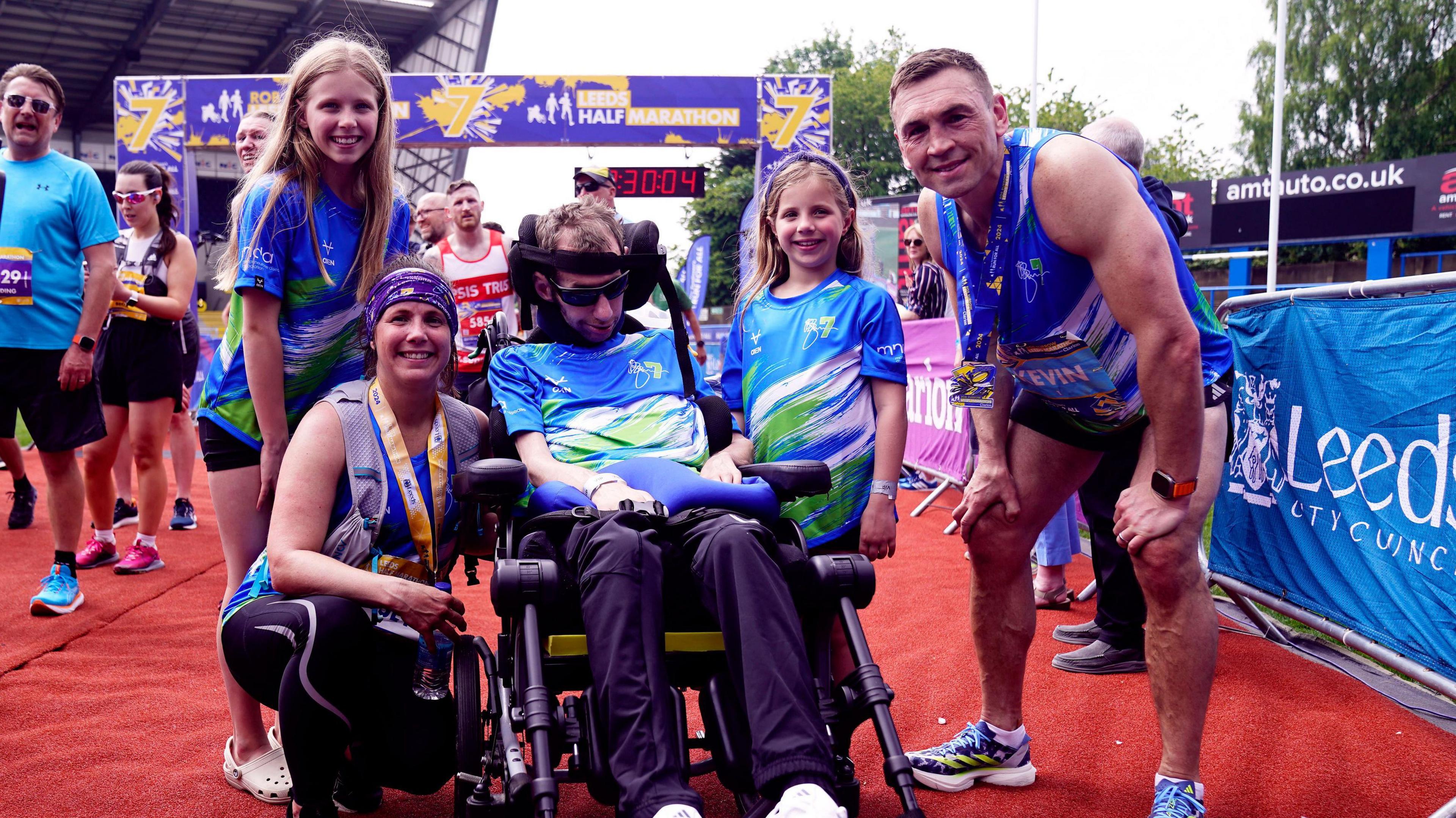 Group picture of Rob Burrow, his wife Lindsey, daughters Macy and Maya and Kevin Sinfield on a red carpet finish line after the Rob Burrow Leeds Marathon.