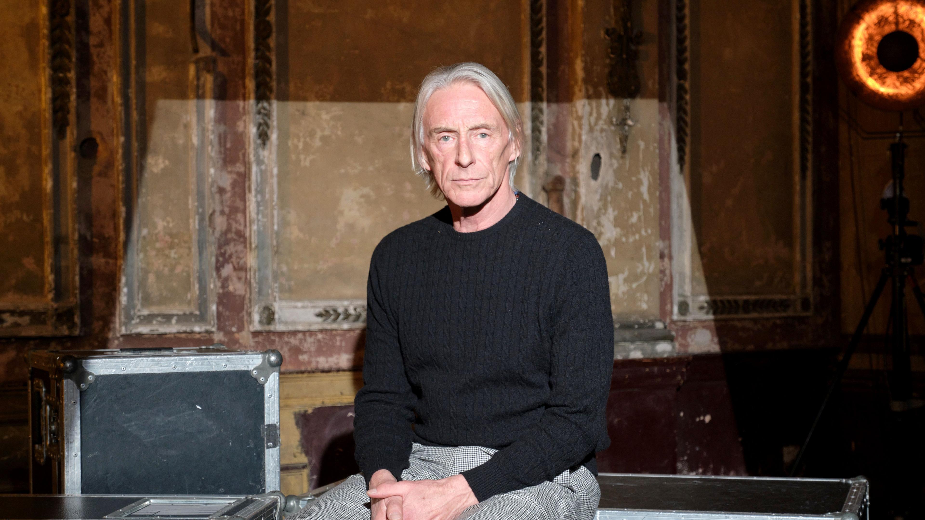 Musician Paul Weller said on a metal music equipment box. He has a black jumper on and grey trousers