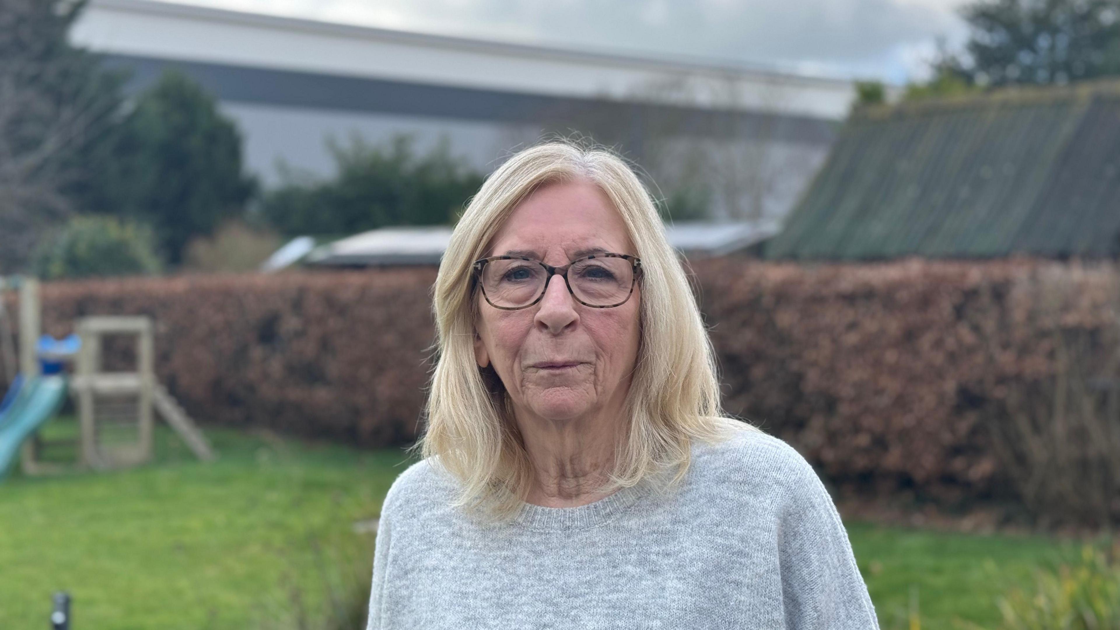 Jenny Upson - a woman wearing a grey jumper standing in a garden. The warehouse can be seen in the background. 