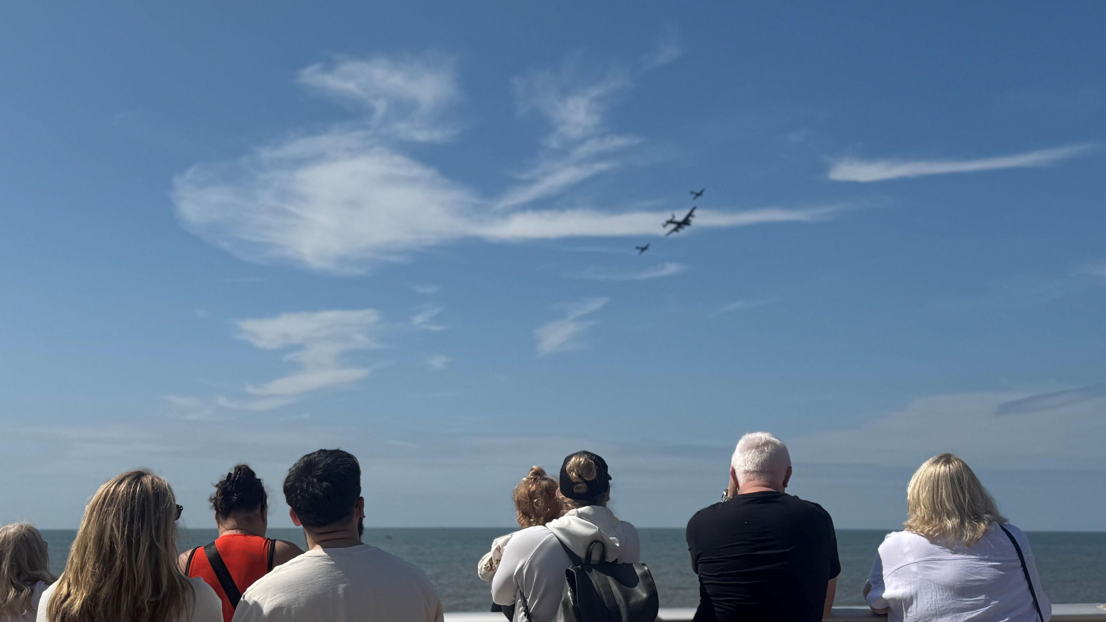 About six people look up at a Battle of Britian memorial flypast, featuring a large military plane flanked by two smaller planes, against wispy white clouds in the blue sky over the sea.