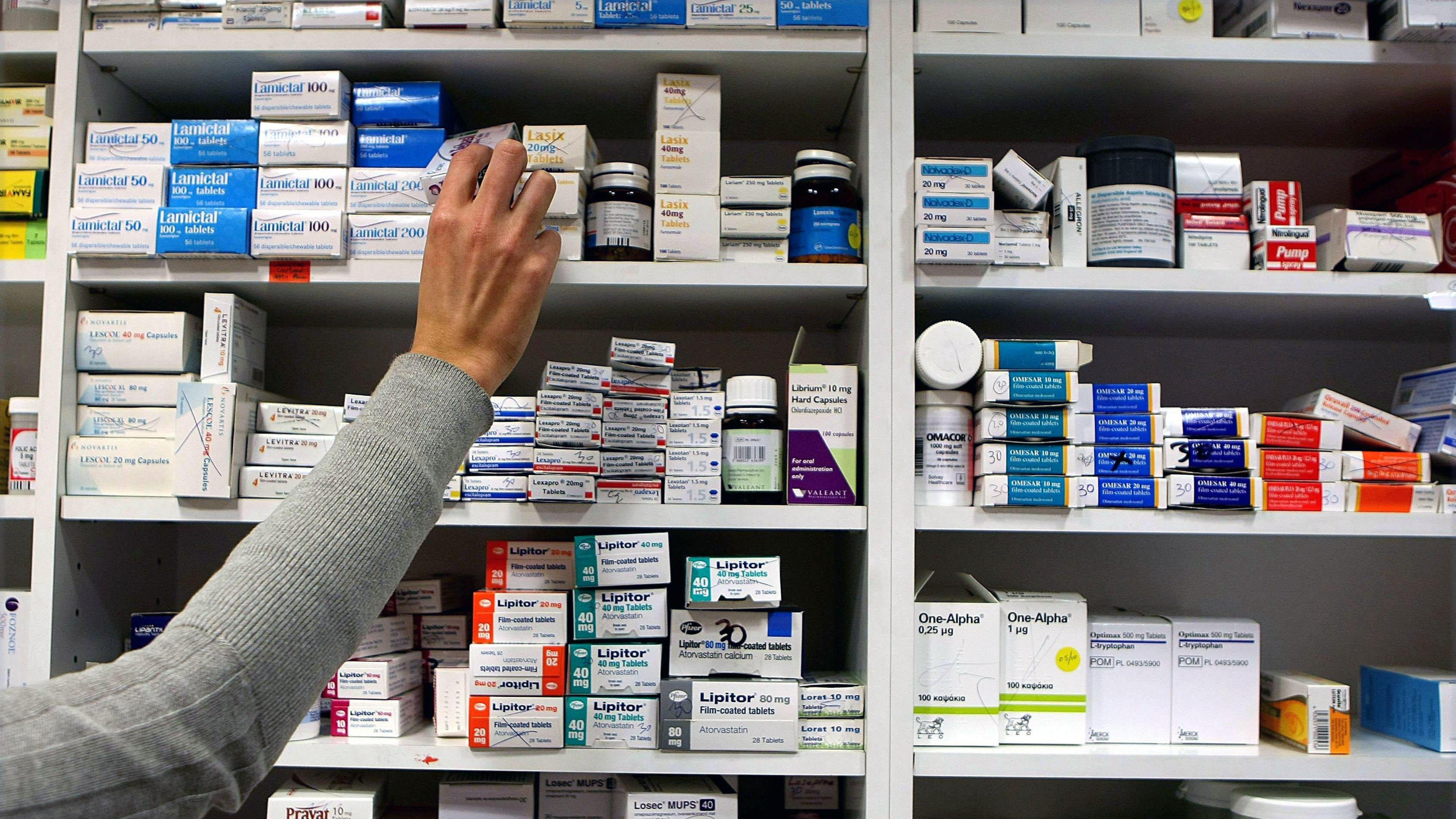 A person in a grey long-sleeve top reaching for a medication box on the top shelf in a pharmacy or medical storage area. The shelves are filled with neatly arranged boxes and containers of various medications, organised by type and brand.