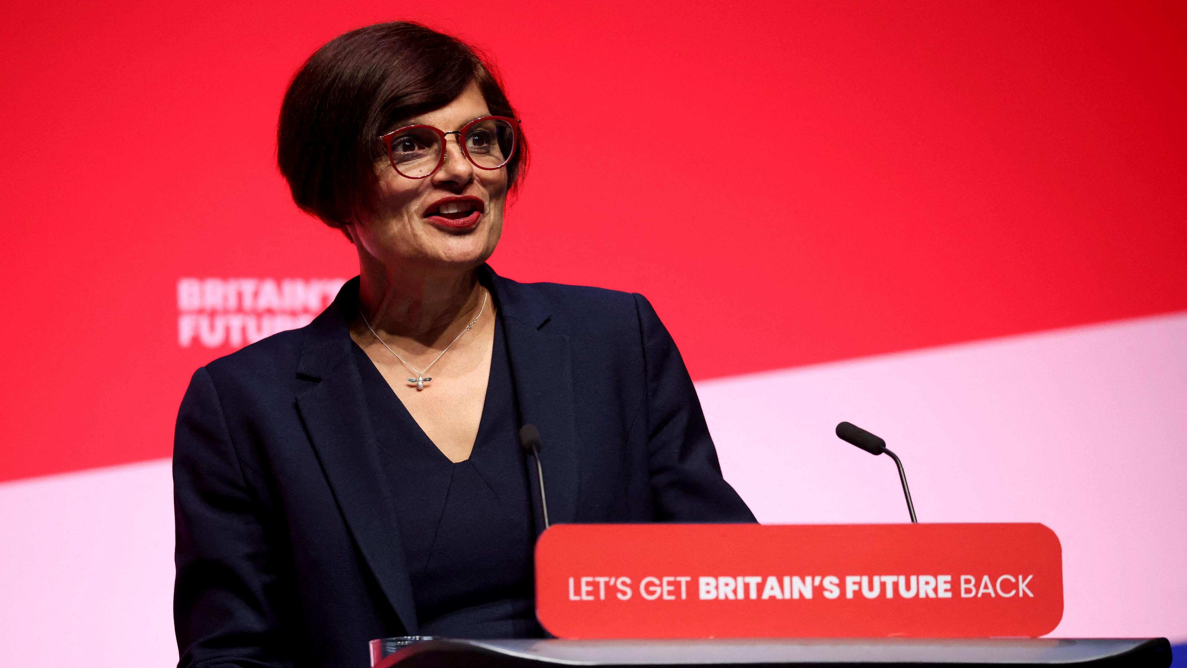 Thangam Debbonaire speaks at a Labour rally, with the podium reading 'Let's Get Britain's Future Back'. She has a red background behind her and wears a black suit.