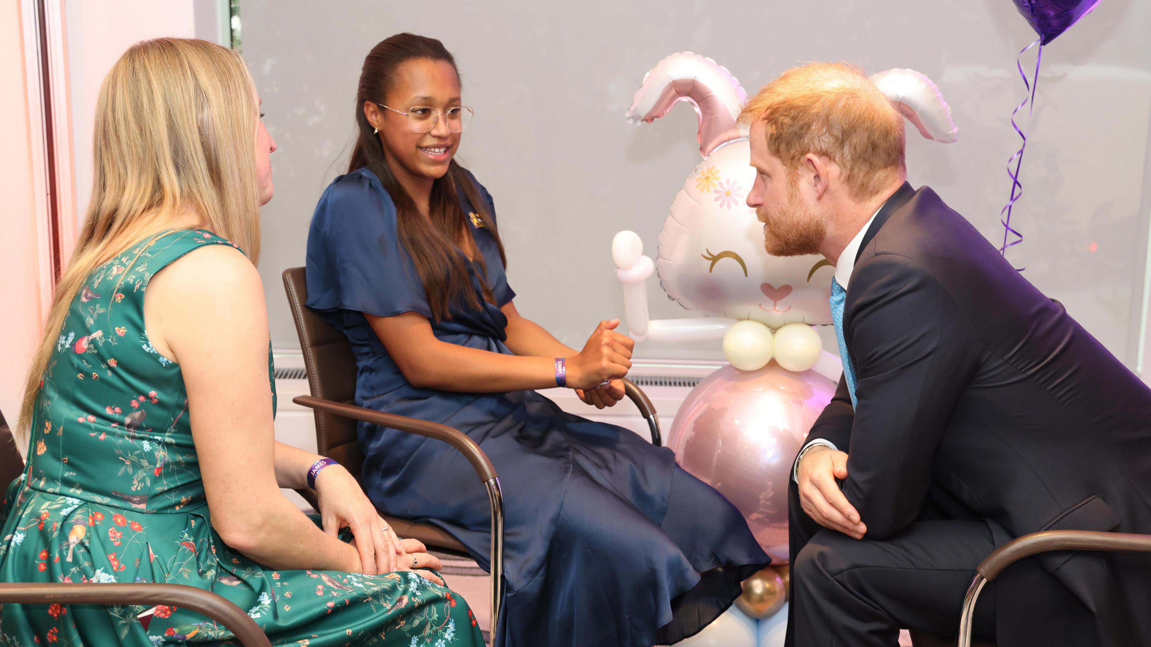 A photograph of Anya (middle) sat in between Prince Harry and her mother Gemma. Anya is wearing glasses and a navy blue dress, while Gemma wears a green dress with flowers on it. Prince Harry wears a dark suit with a white shirt and blue tie