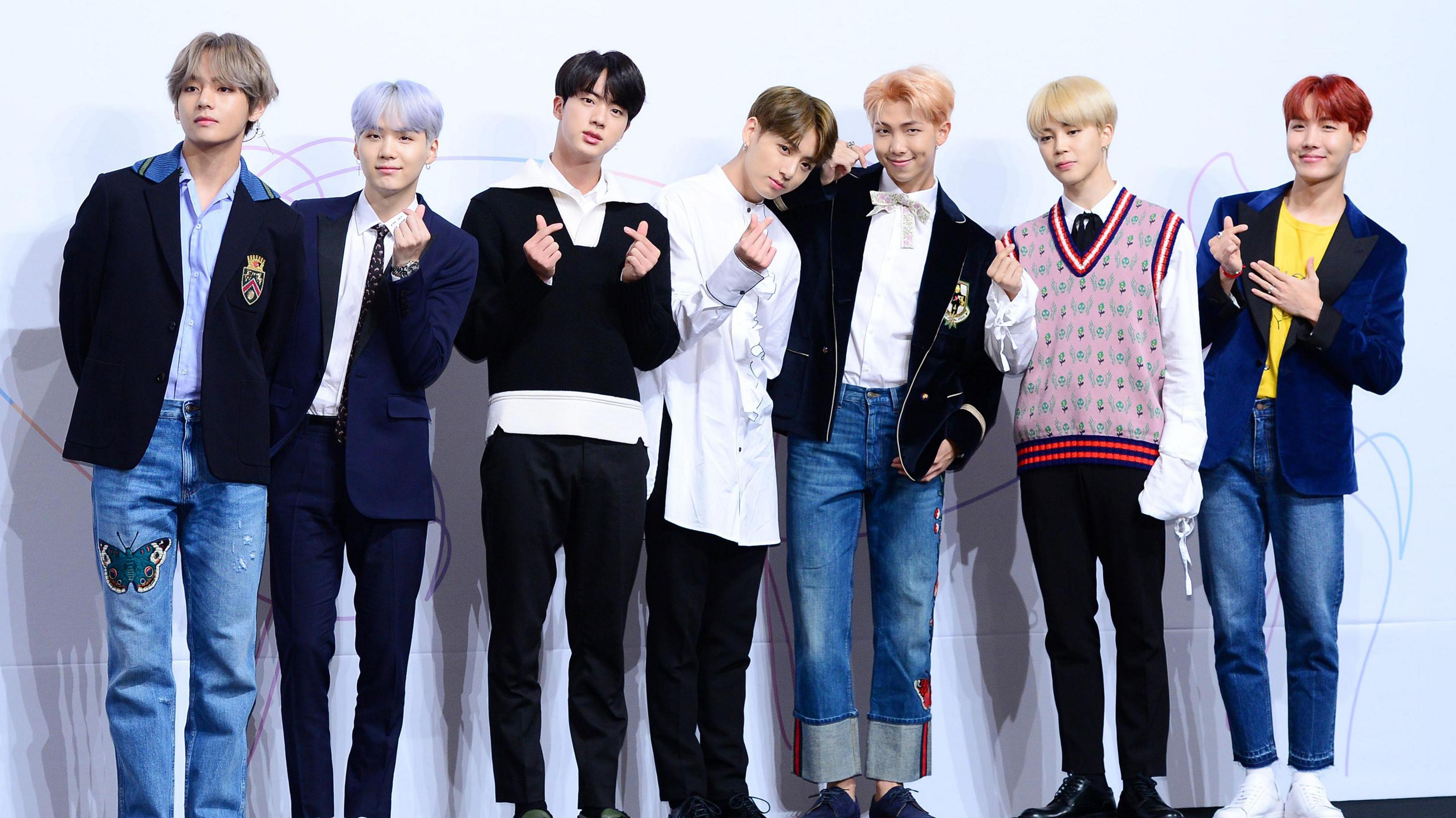 From left to right, V wears blue jeans with a butterfly on the thigh and a dark suit jacket, he has light silvery hair. Suga wears a navy suit with a tie, he has light purple hair. Jin wears black trousers and a black sweater with white shirt, he has black hair. Jung Kook wears a long white shirt and black trousers, he has brown hair. RM has a white shirt and black suit jacket with jeans, he has light orange hair. Jimin wears a pink, red and navy sweater vest with white shirt and black trousers, he has blond hair. J-Hope has jeans and a yellow t-shirt with navy jacket on, he has ginger hair. 