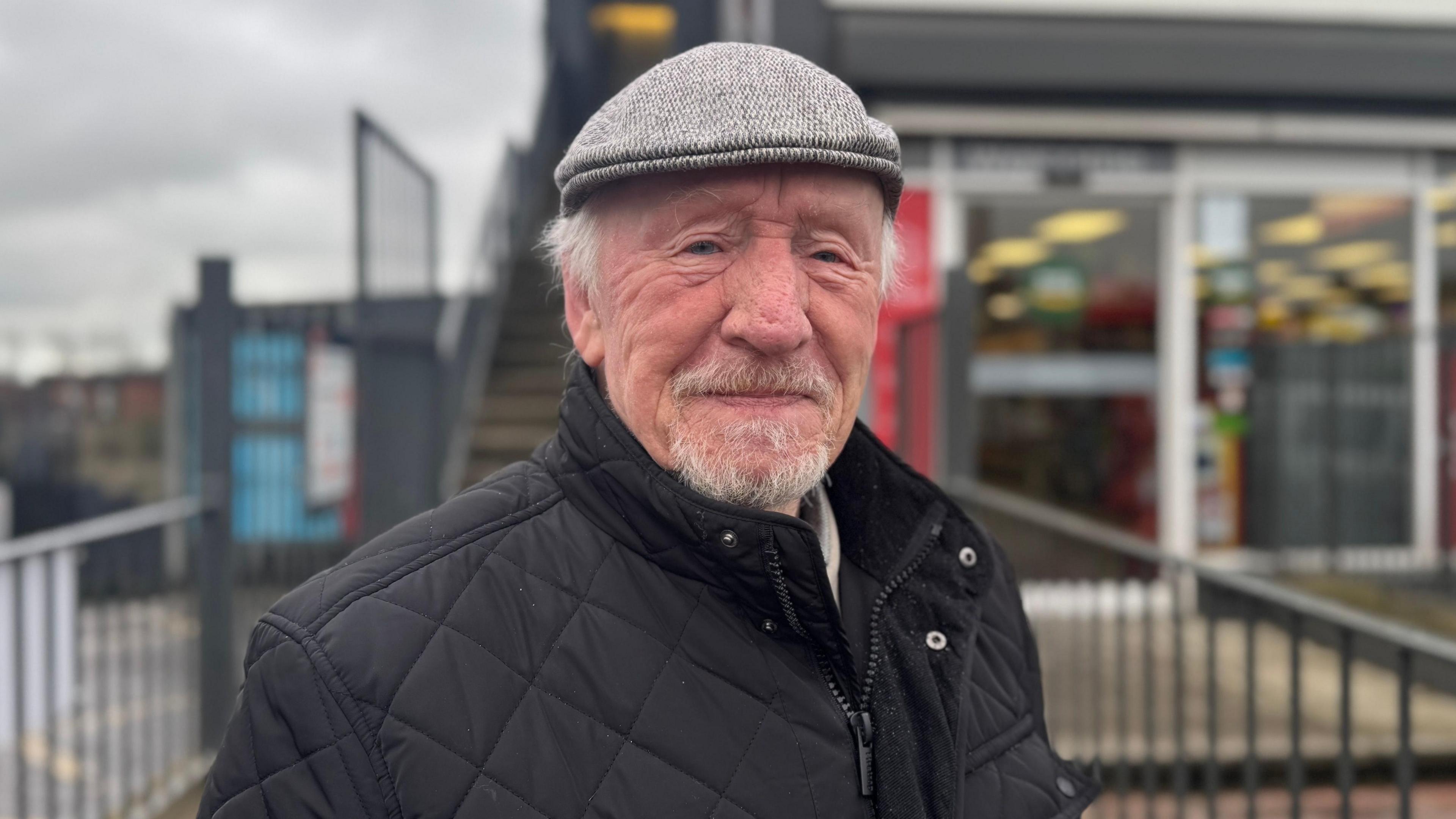 Roy Fallis. A man wears a grey flat cap and is smiling at the camera. He has white hair and a white moustache and goatie. He is wearing a black, padded raincoat and is standing in front of a local shop in Carryduff.