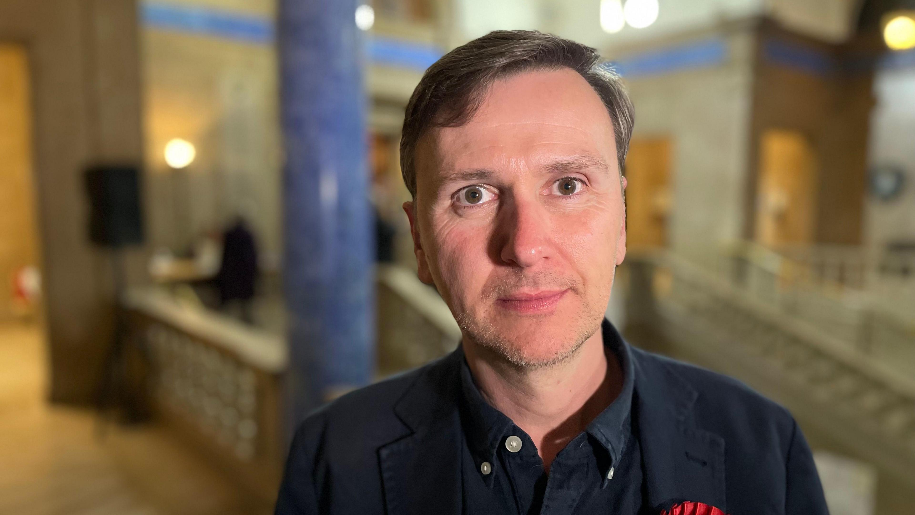 Andrew wearing a black shirt and blazer with a red rosette- partially showing 